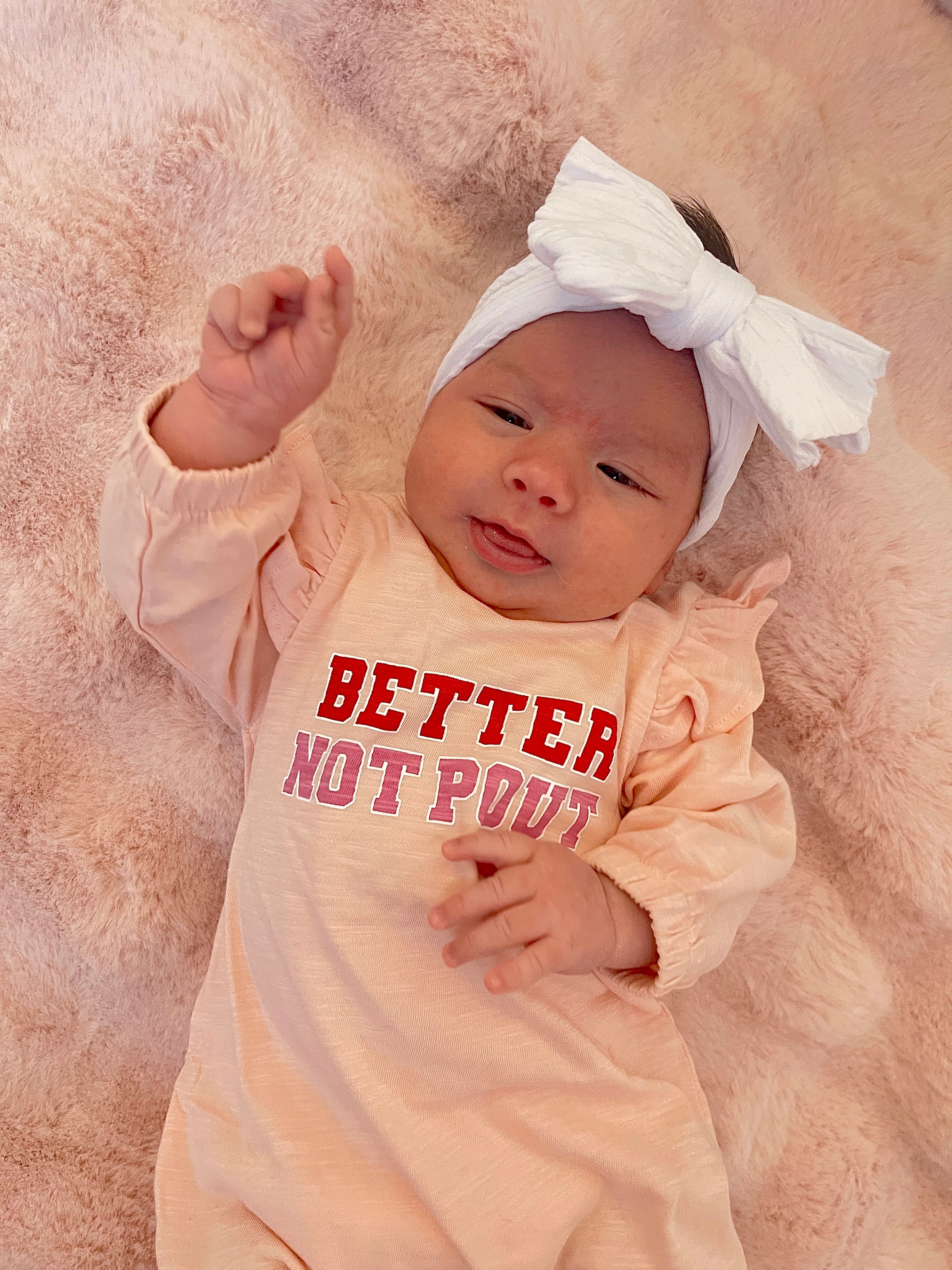 Smiling baby wearing a "Better Not Pout" outfit with a large white bow, resting on a soft pink blanket.