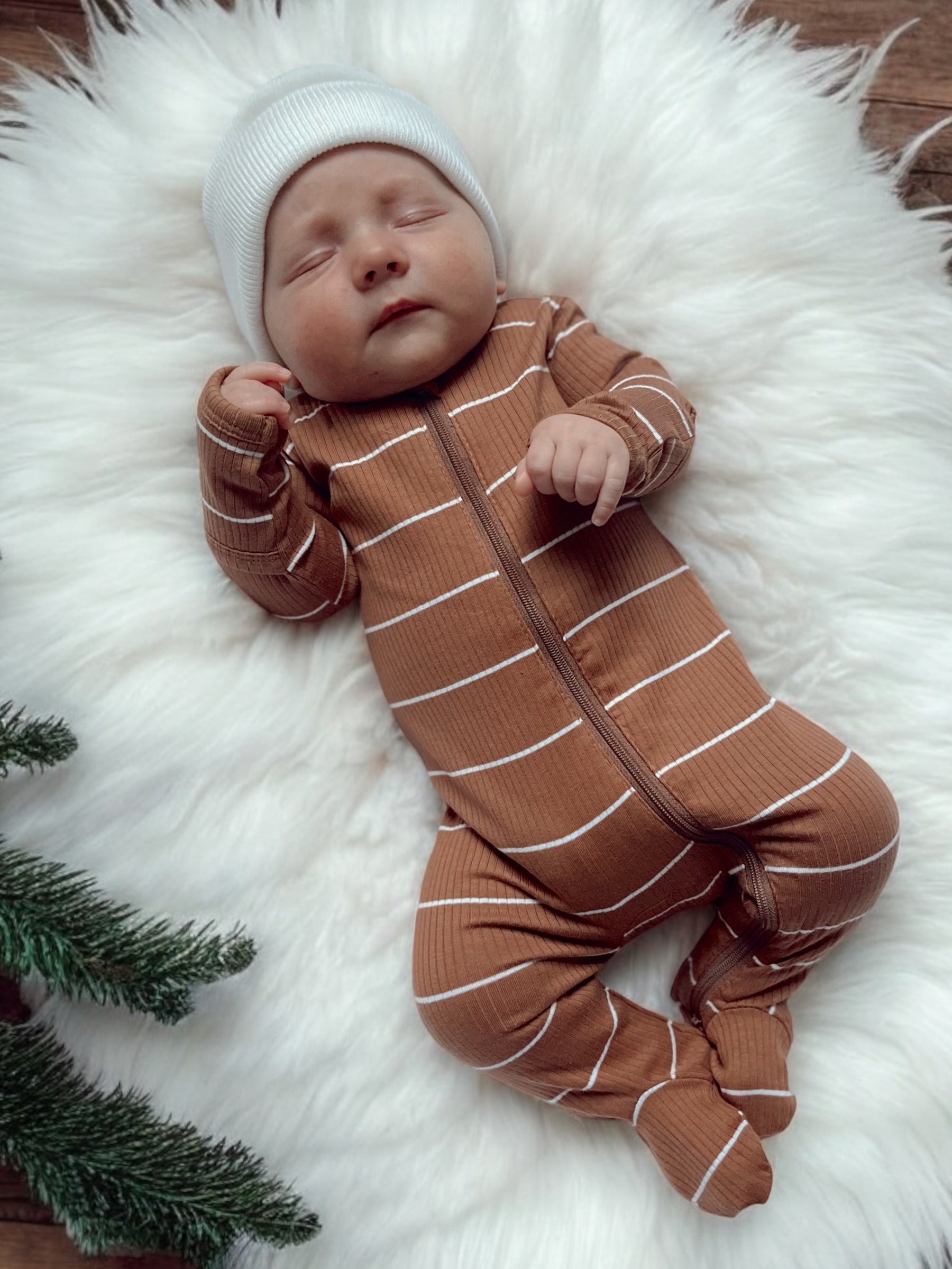 Sleeping baby in a brown striped onesie on a fluffy white rug with greenery nearby.