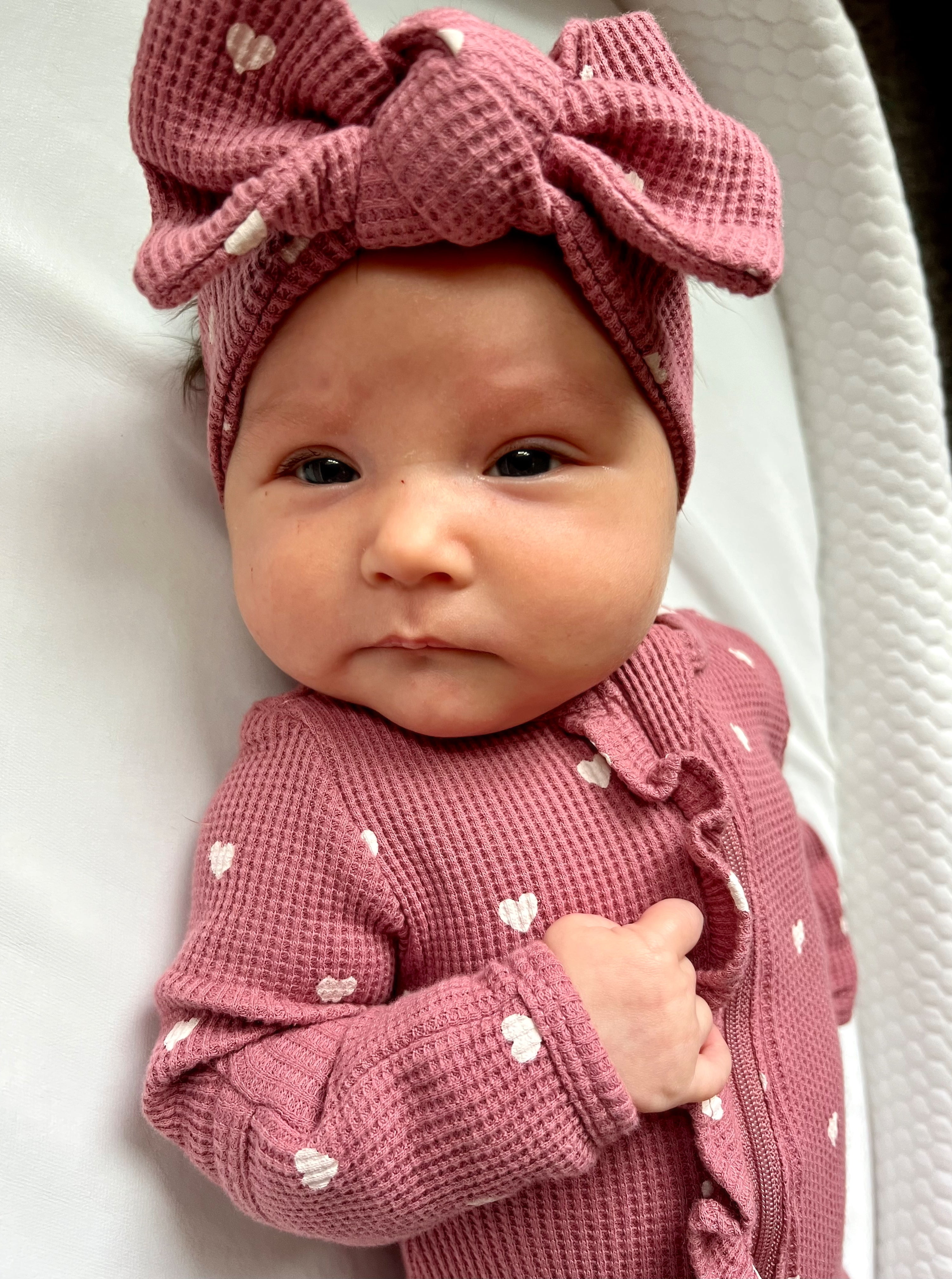 Baby with a pink bow headband and heart-patterned outfit, laying on a white surface, looking at the camera.