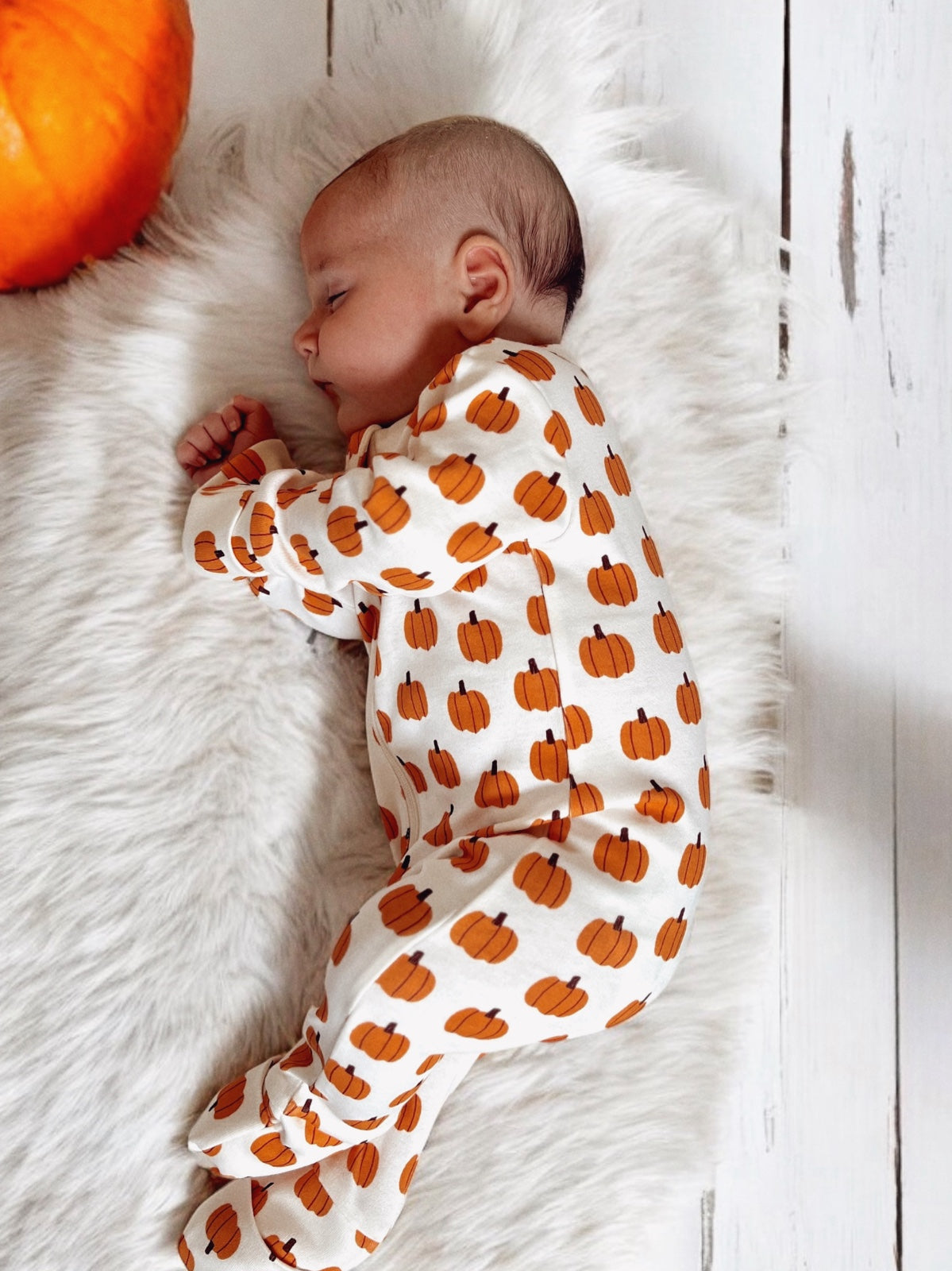 Sleeping baby in pumpkin-patterned pajamas on a soft faux fur blanket, with a pumpkin nearby.