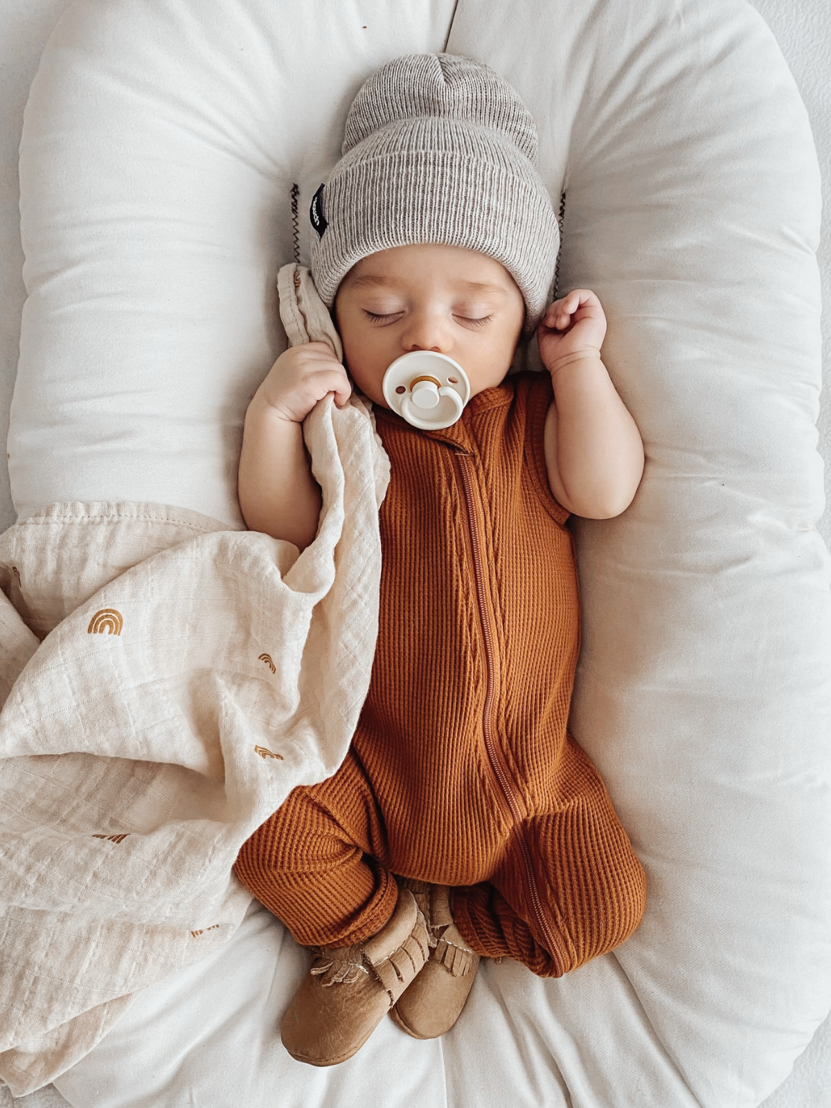 Infant sleeping peacefully in a cozy environment, wearing a beanie and stylish outfit, with a pacifier and blanket.