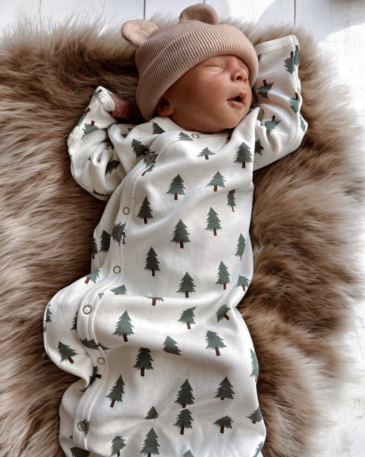 Sleeping baby in a bear-ear hat, dressed in a tree-printed outfit on a soft, furry blanket.