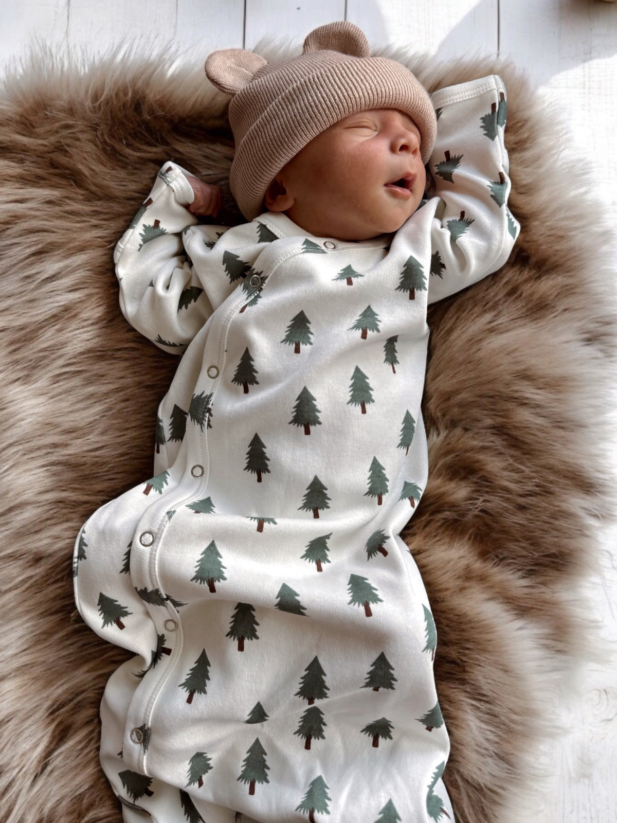 Sleeping baby in a bear-ear hat, dressed in a tree-printed outfit on a soft, furry blanket.