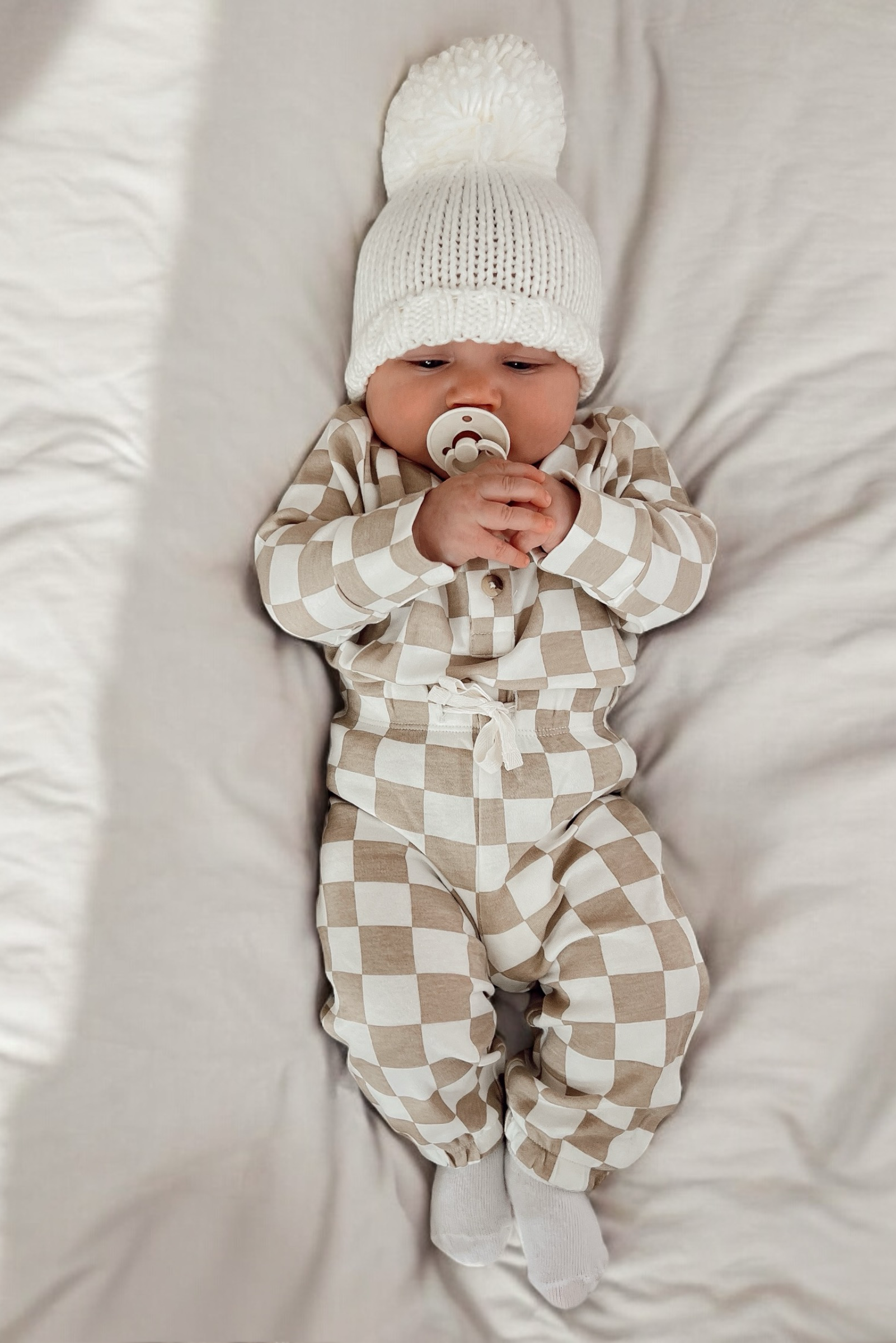 Baby lying on a blanket, wearing a white pom-pom hat and beige checkered outfit, holding a pacifier.