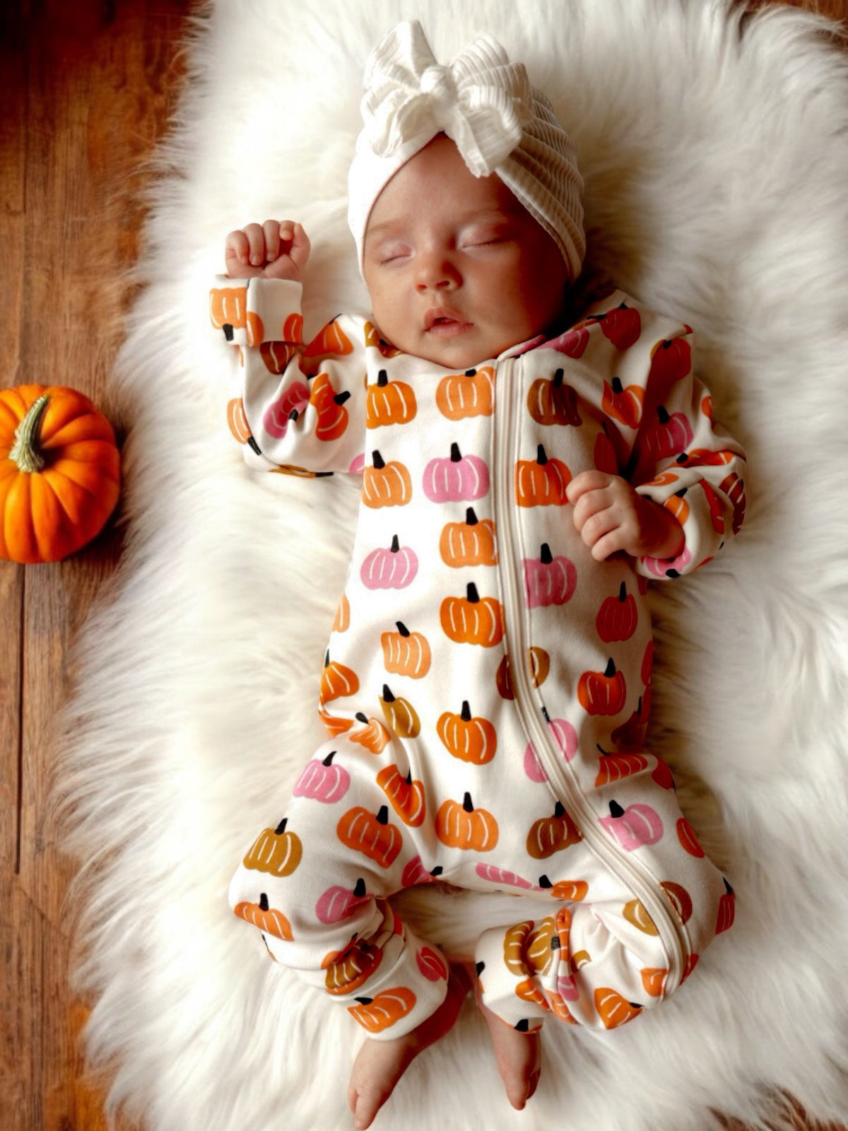 Baby sleeping on fluffy blanket, wearing a pumpkin-patterned outfit, with a small pumpkin nearby.