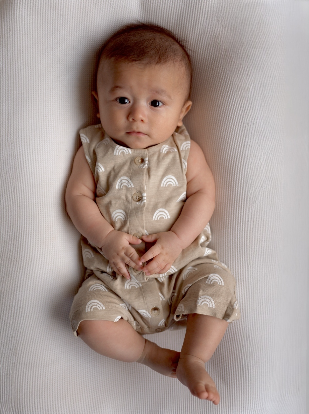A baby with short hair, wearing a beige outfit with rainbow patterns, sitting on a textured white surface.