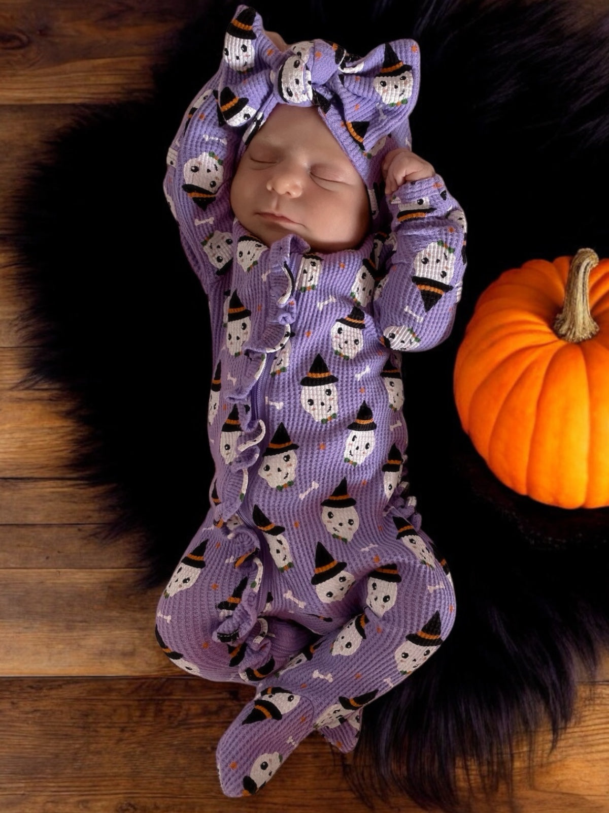 Baby in purple Halloween-themed outfit with ghost print, lying on dark fur rug next to a pumpkin.