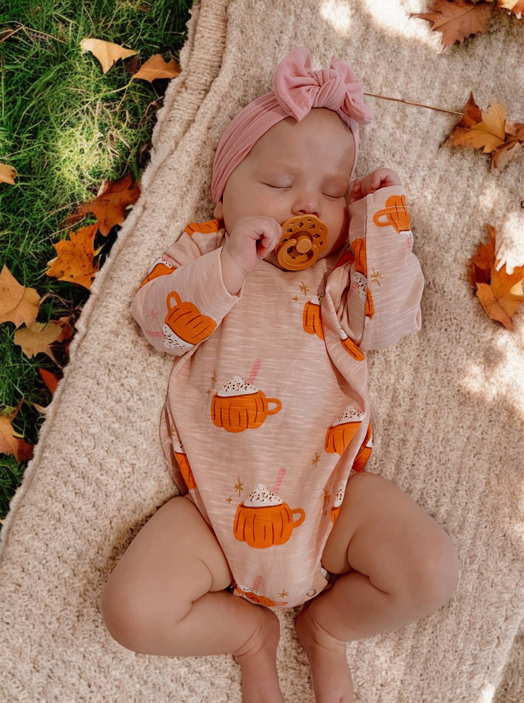 Infant sleeping on a blanket, wearing a peach bodysuit with mug graphics and a pink headband, surrounded by autumn leaves.