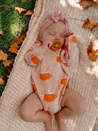 Infant sleeping on a blanket, wearing a peach bodysuit with mug graphics and a pink headband, surrounded by autumn leaves.