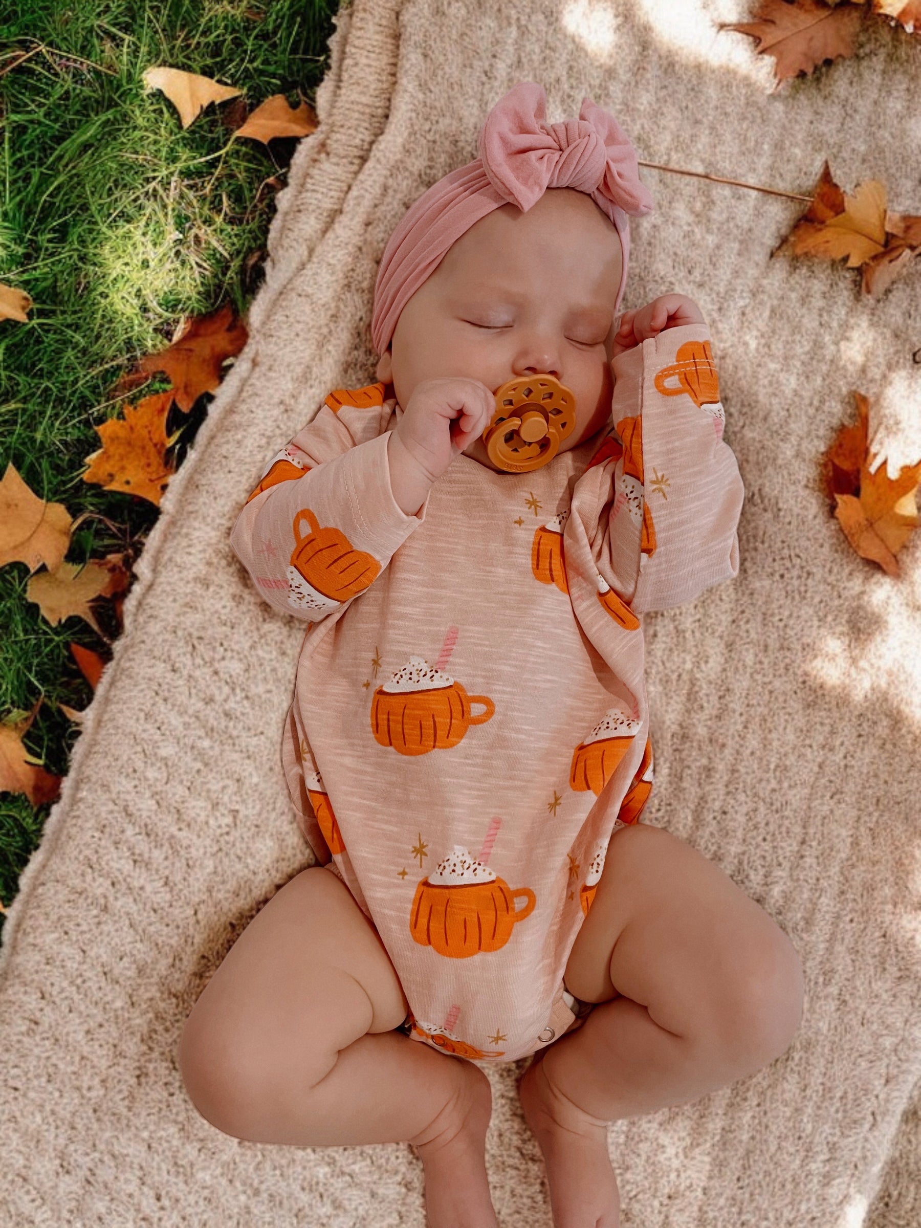 Infant sleeping on a blanket, wearing a peach bodysuit with mug graphics and a pink headband, surrounded by autumn leaves.