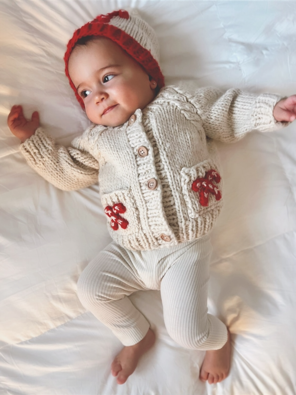 Smiling baby lying on a white surface, wearing a knit cream sweater with red accents and matching hat.