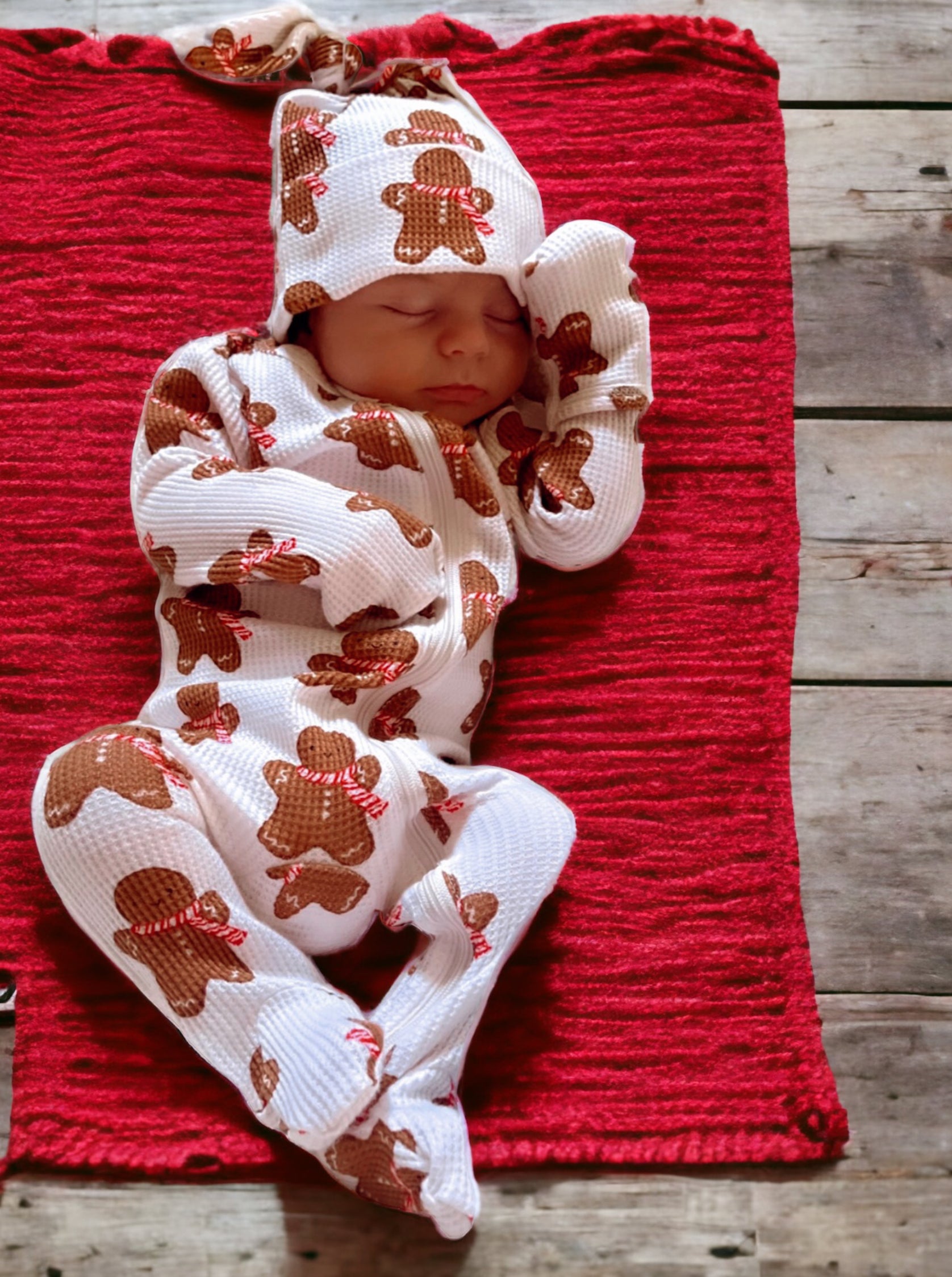 Baby sleeping peacefully on a red blanket, wearing a white outfit with gingerbread patterns.