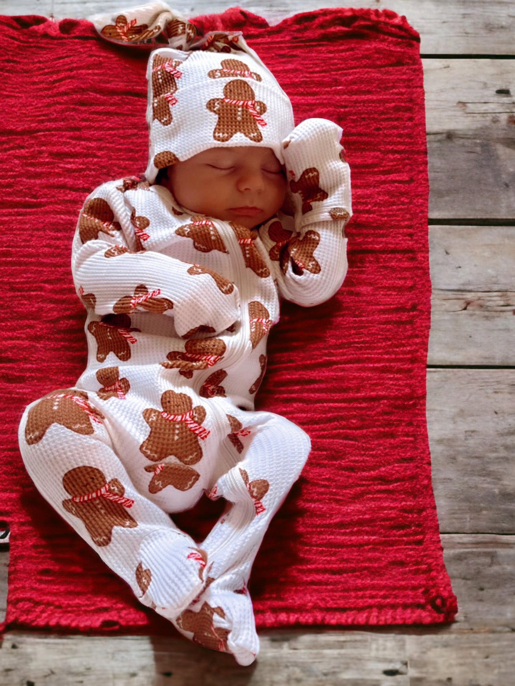 Baby sleeping peacefully on a red blanket, wearing a white outfit with gingerbread patterns.
