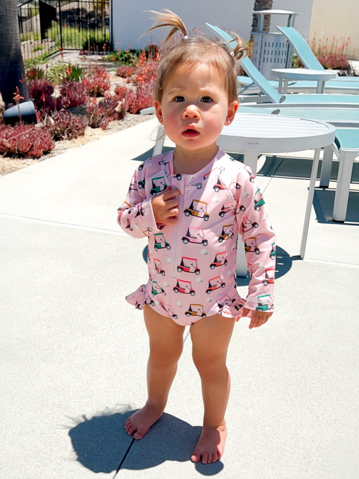 Toddler in a pink golf cart-patterned swimsuit stands on a sunny patio with outdoor furniture and landscaping.