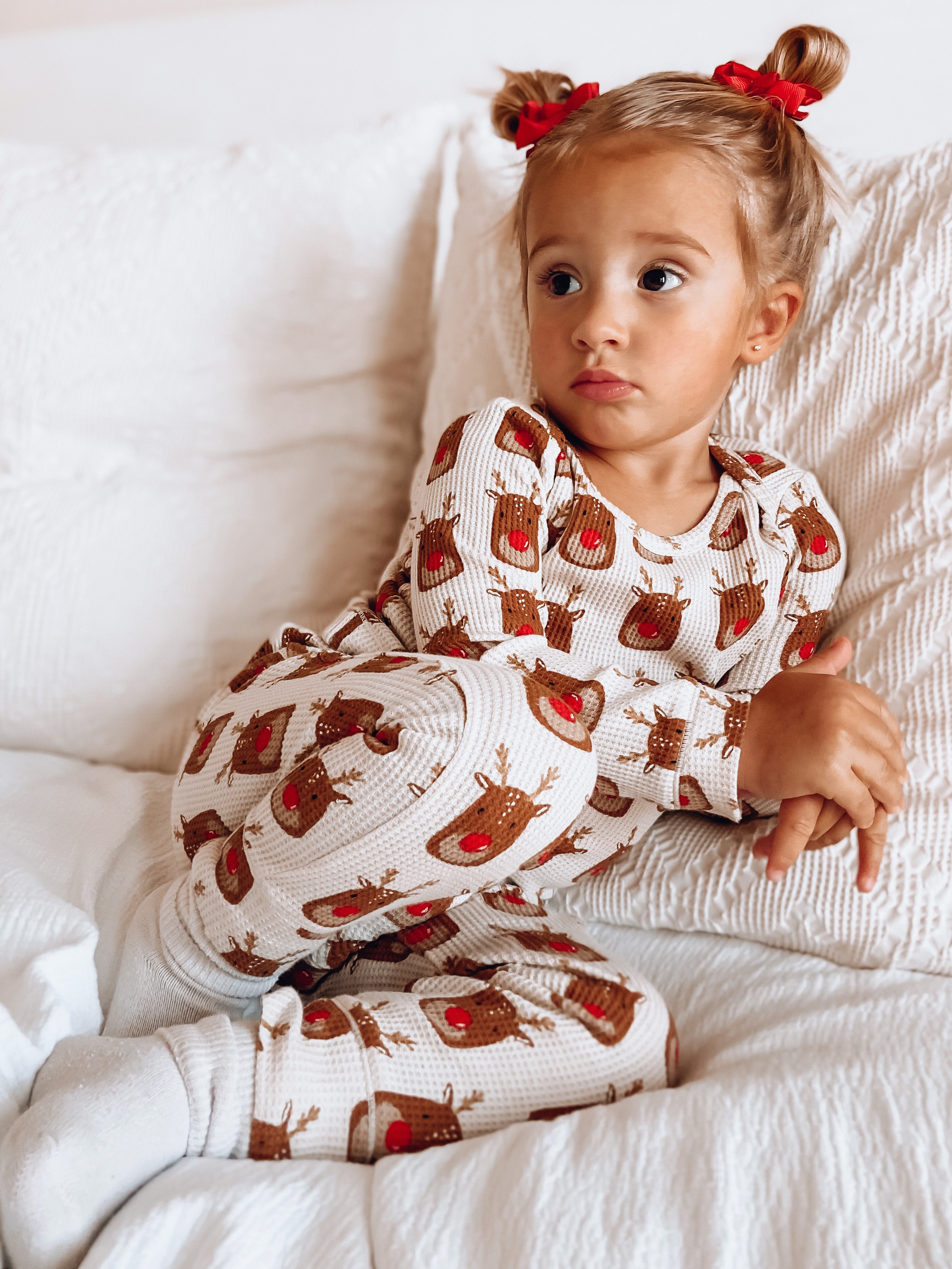 Toddlers in festive reindeer pajamas sitting on a couch, looking thoughtful with hair in two buns.