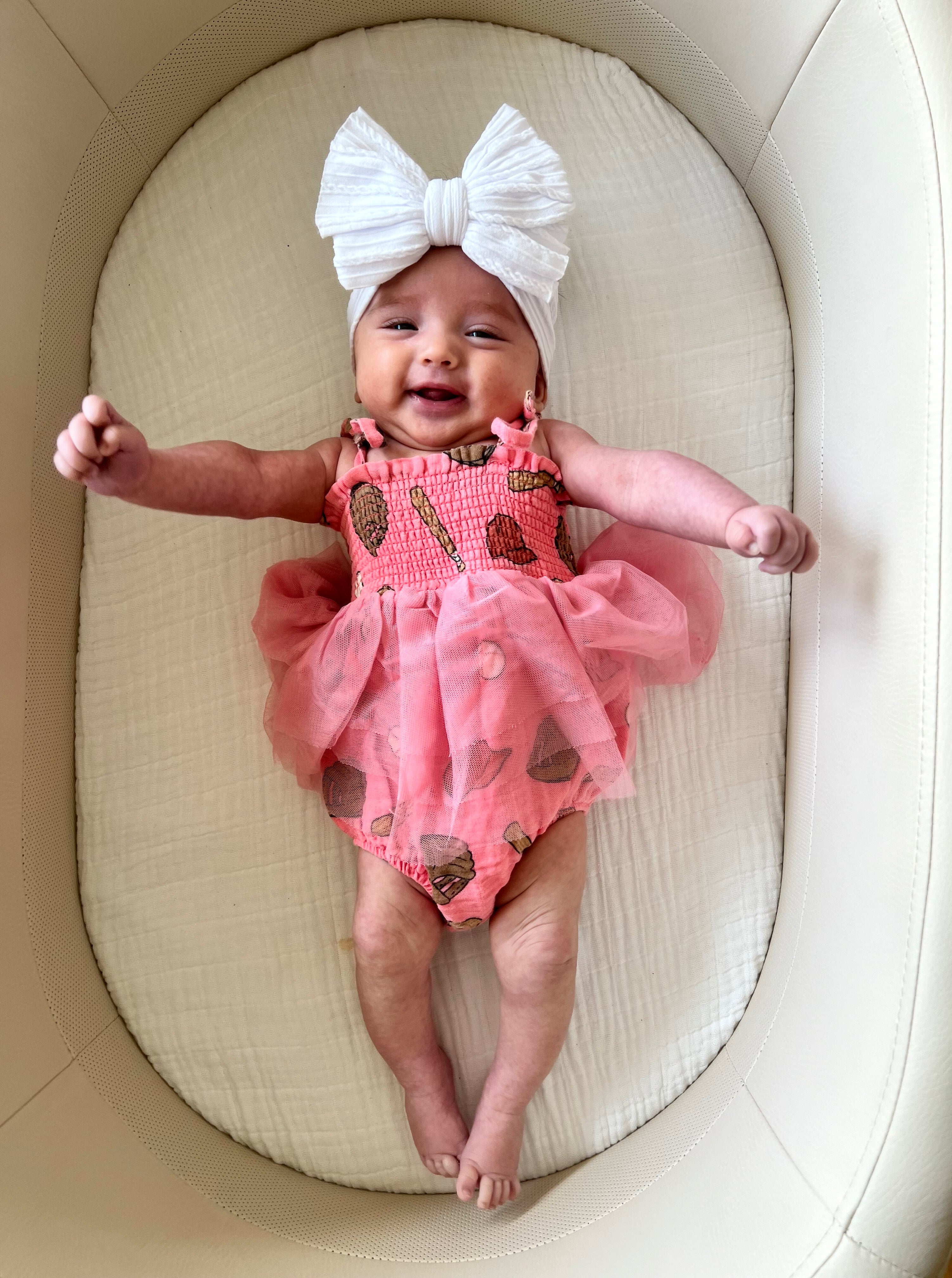 Smiling baby in a pink dress with a large white bow, lying on a soft, neutral-colored surface.