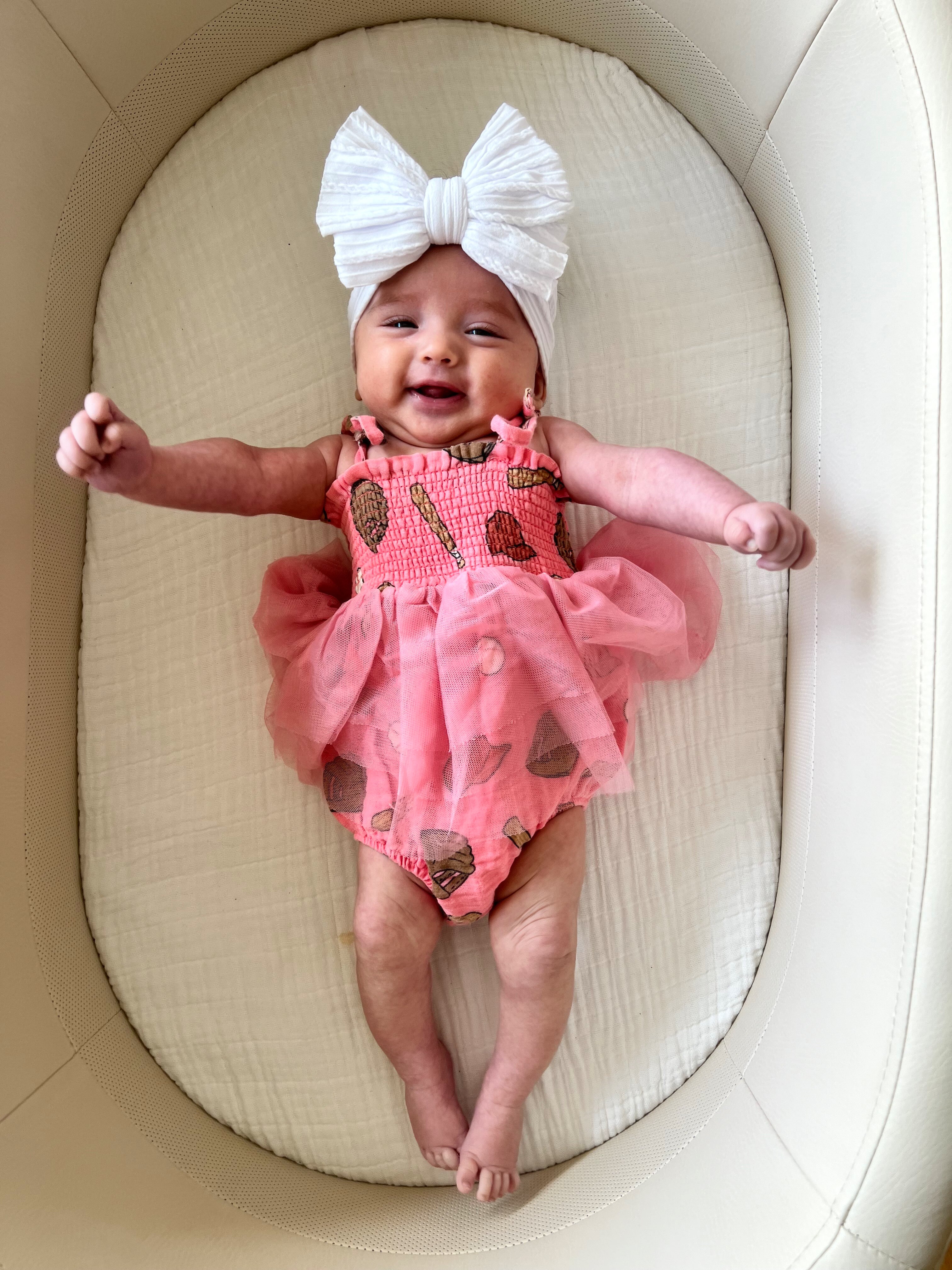 Smiling baby in a pink dress with a large white bow, lying on a soft, neutral-colored surface.
