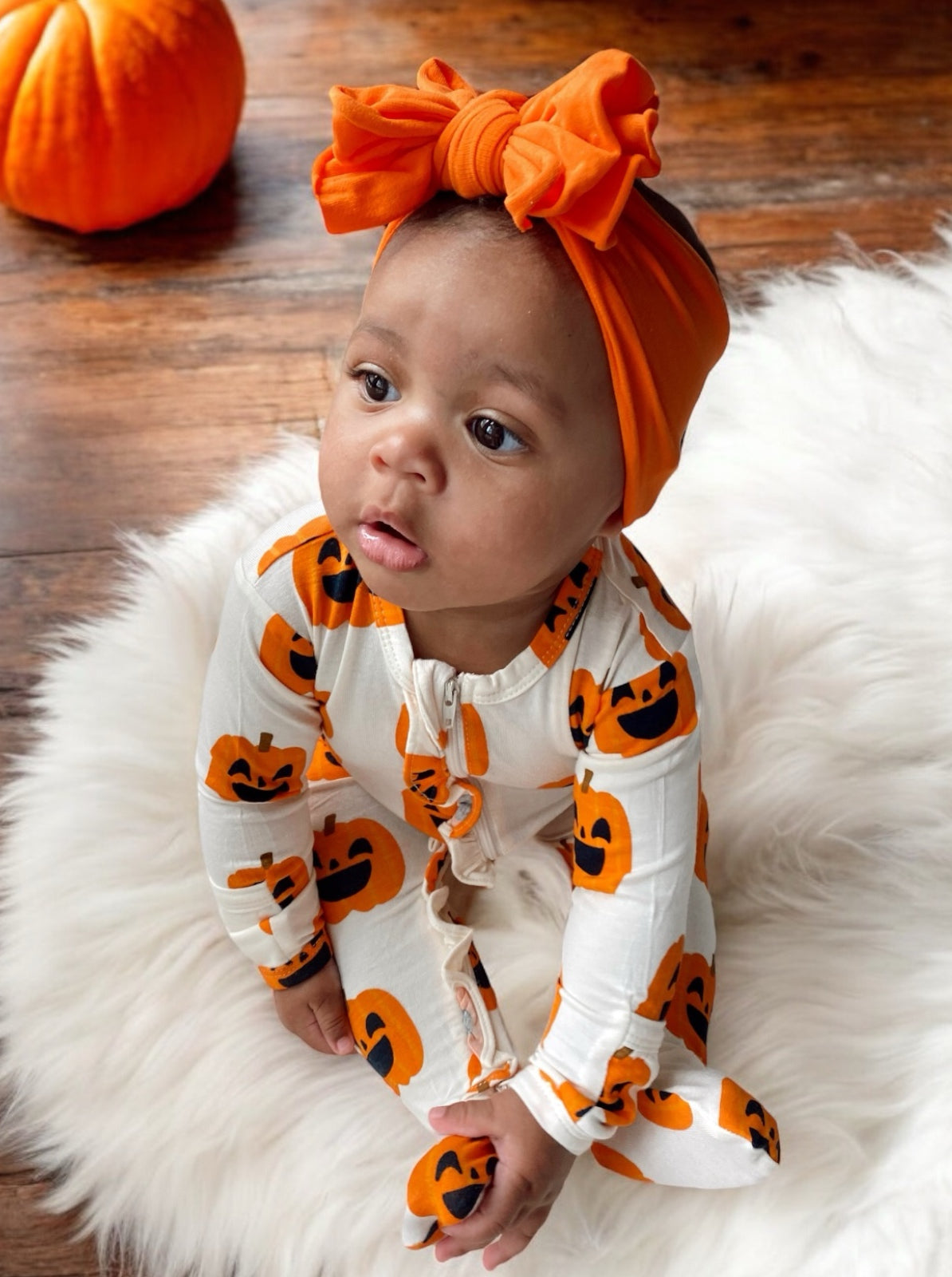 Baby wearing pumpkin-themed pajamas and an orange headband sits on a fluffy rug, with a pumpkin nearby.