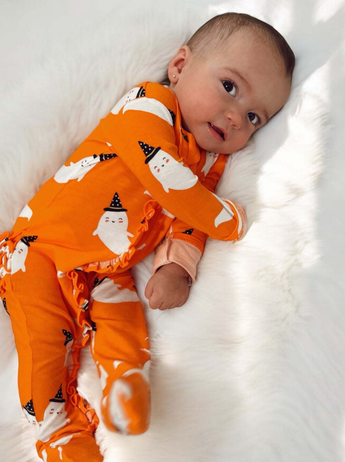 Baby in orange ghost print pajamas lying on a white fur blanket, smiling and looking at the camera.