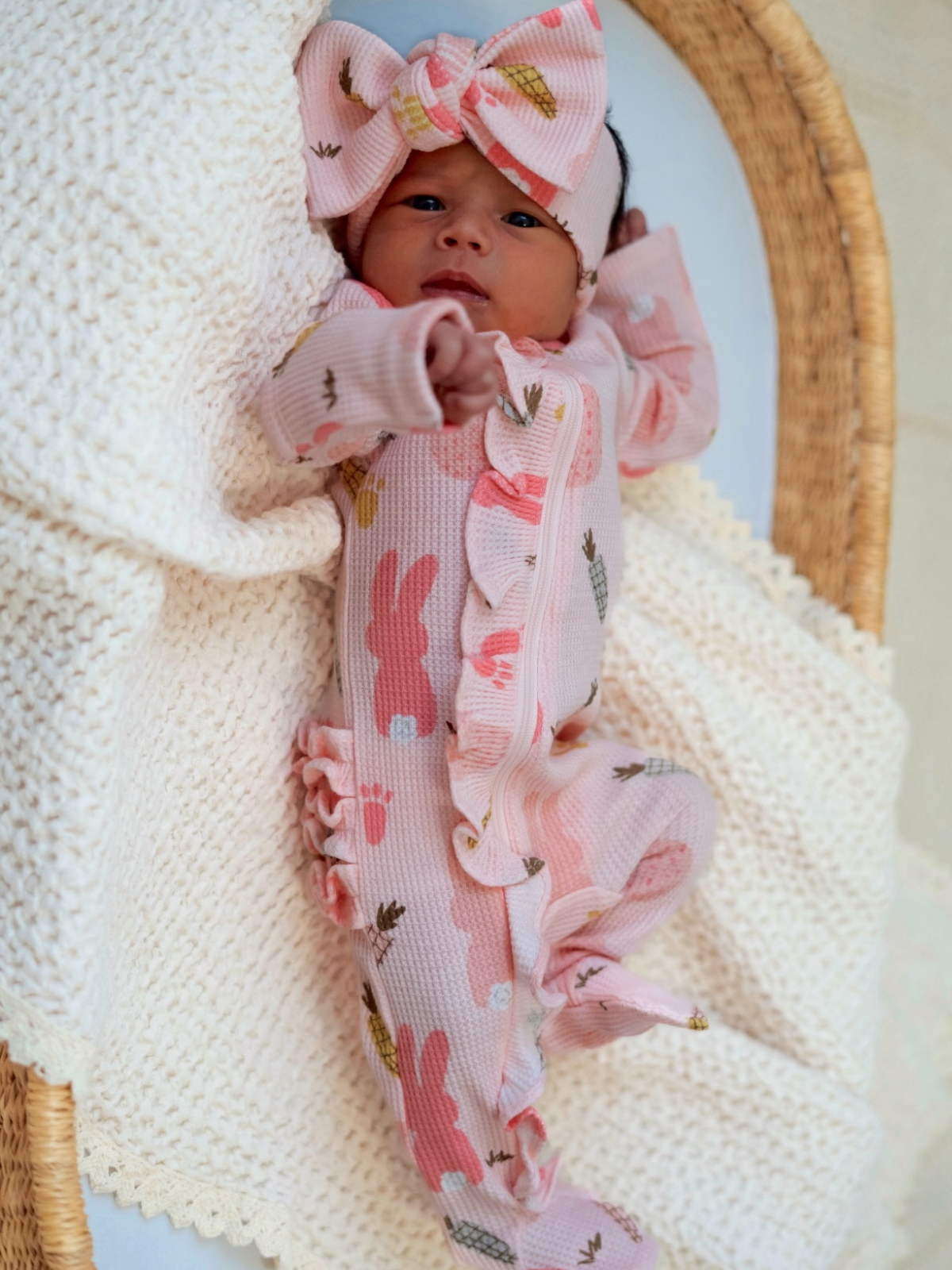 Newborn baby in pink floral outfit with a matching bow, resting on a knitted blanket in a wicker basket.