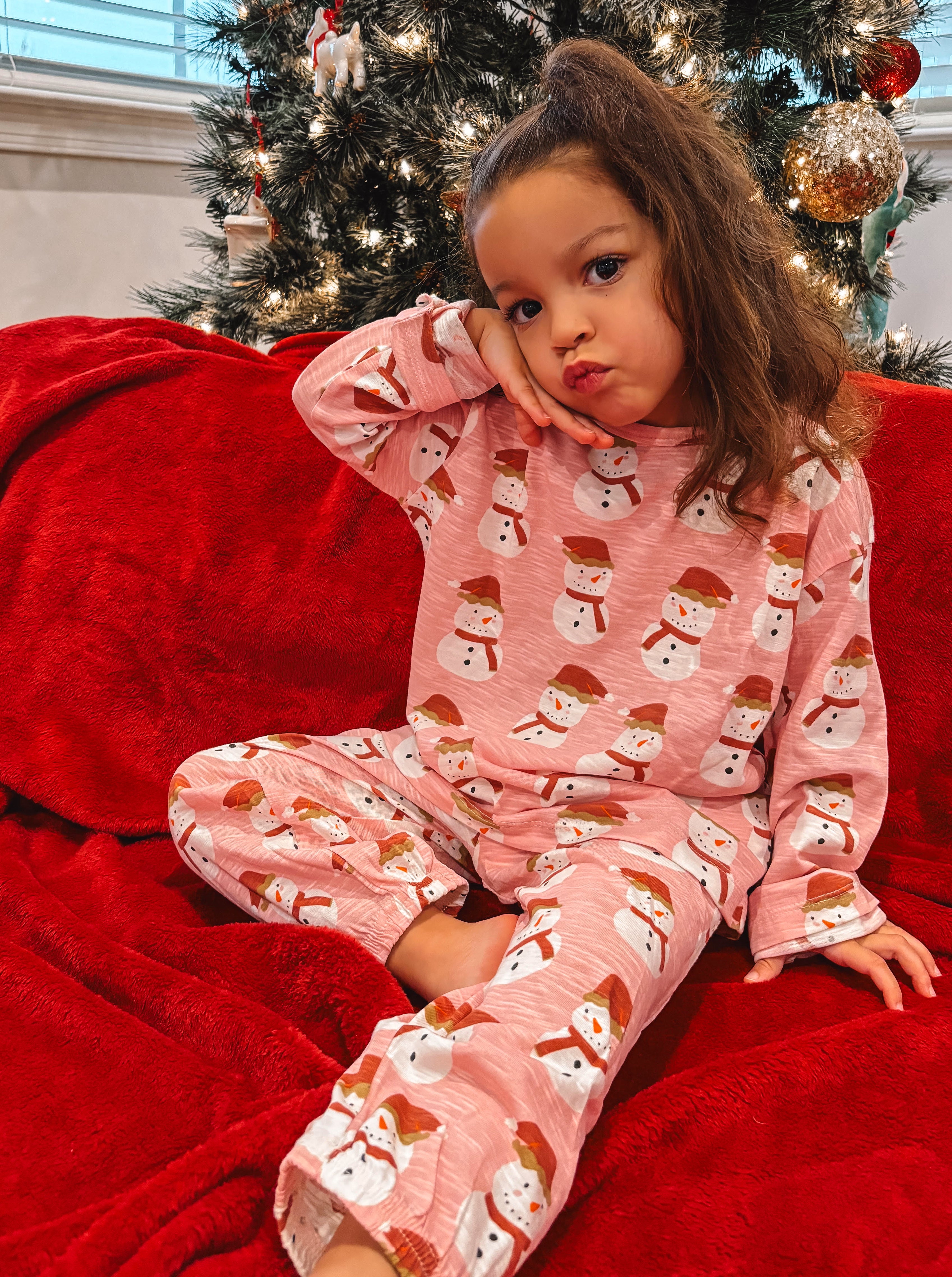 Young girl in festive snowman pajamas posing on a red blanket in front of a Christmas tree.