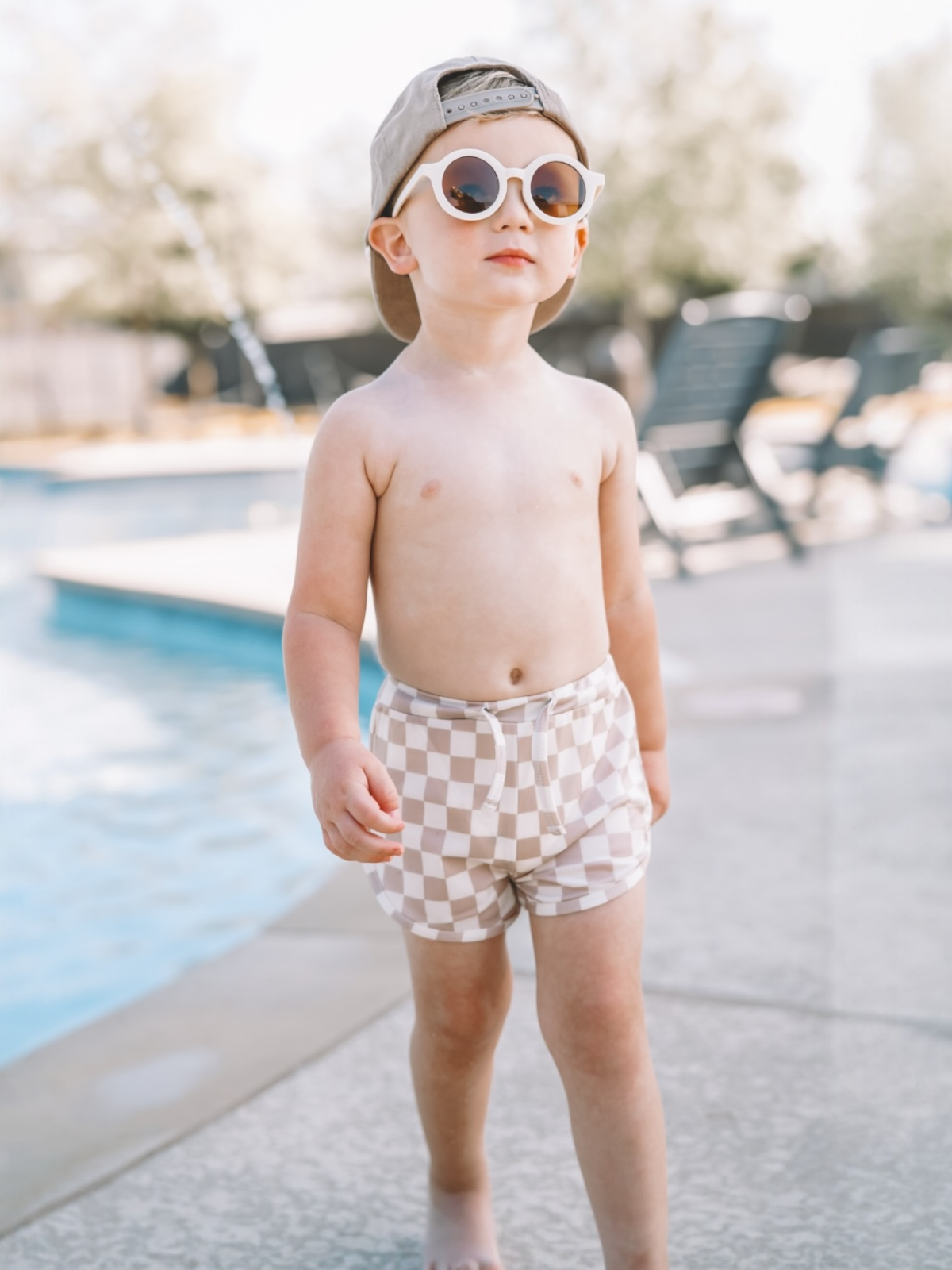 Child in checkered swim shorts and sunglasses walking by a pool on a sunny day.