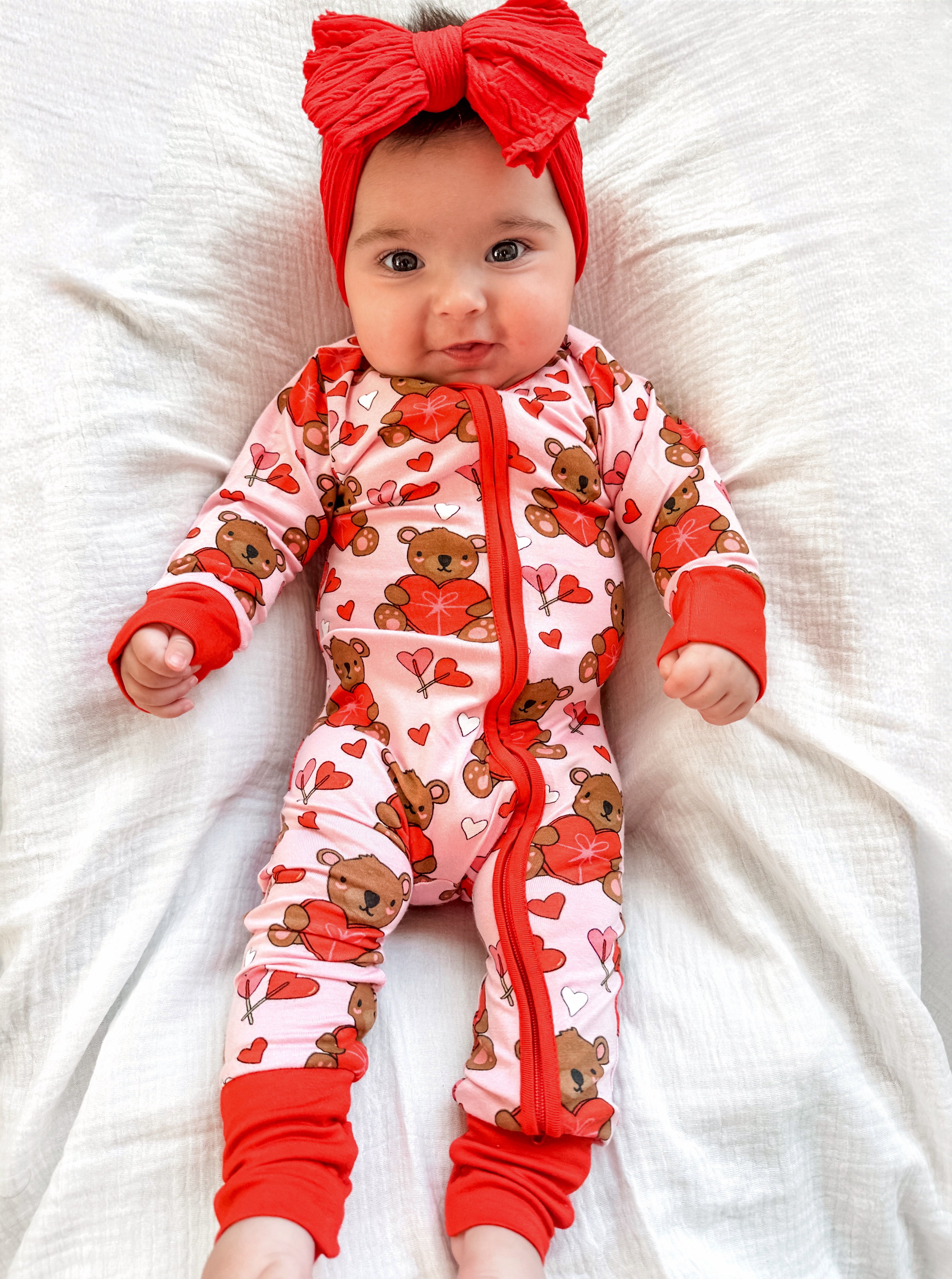 Baby in a pink teddy bear onesie with hearts, wearing a red headband, lying on a white blanket.