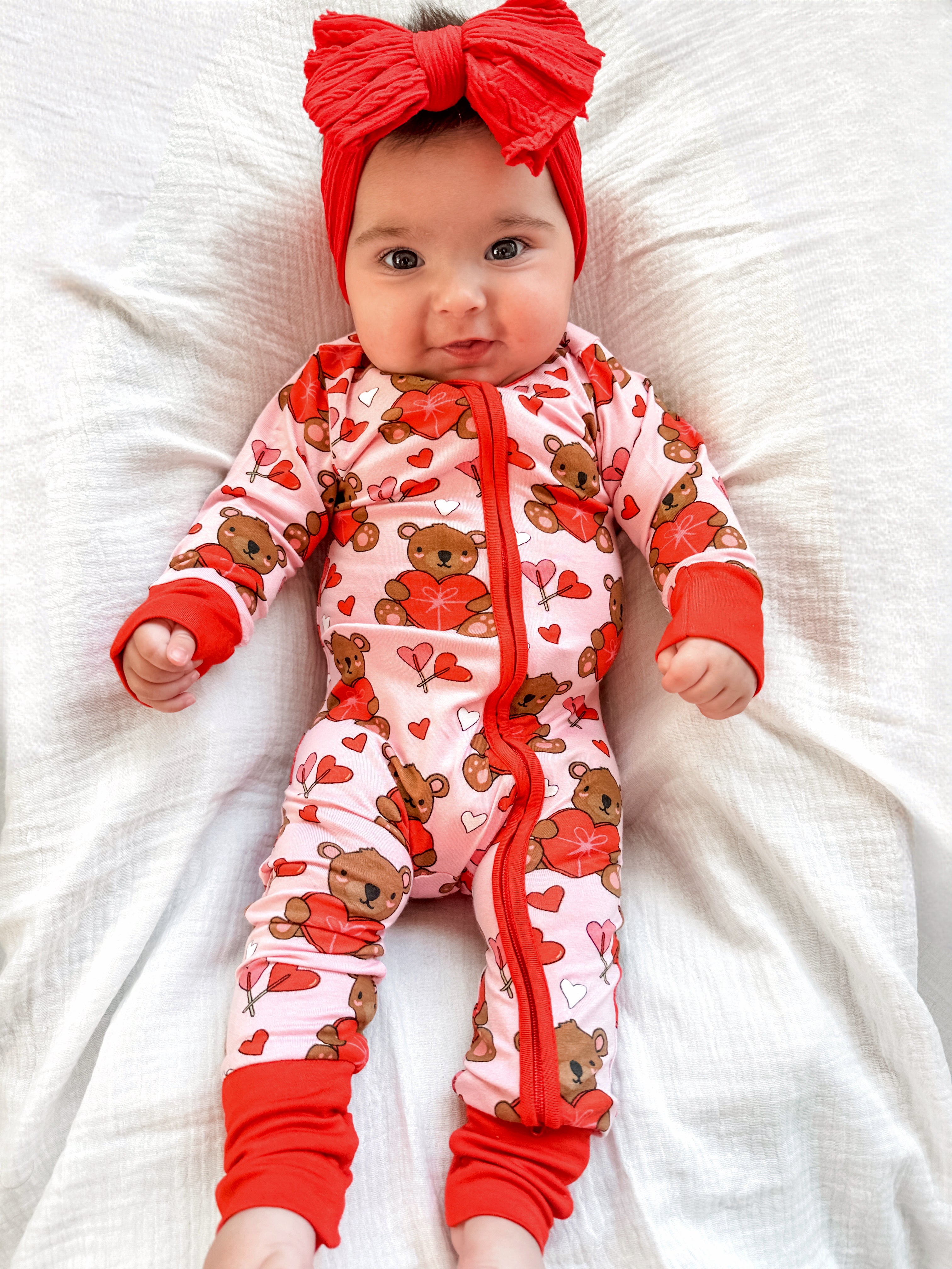 Baby in a pink teddy bear onesie with hearts, wearing a red headband, lying on a white blanket.