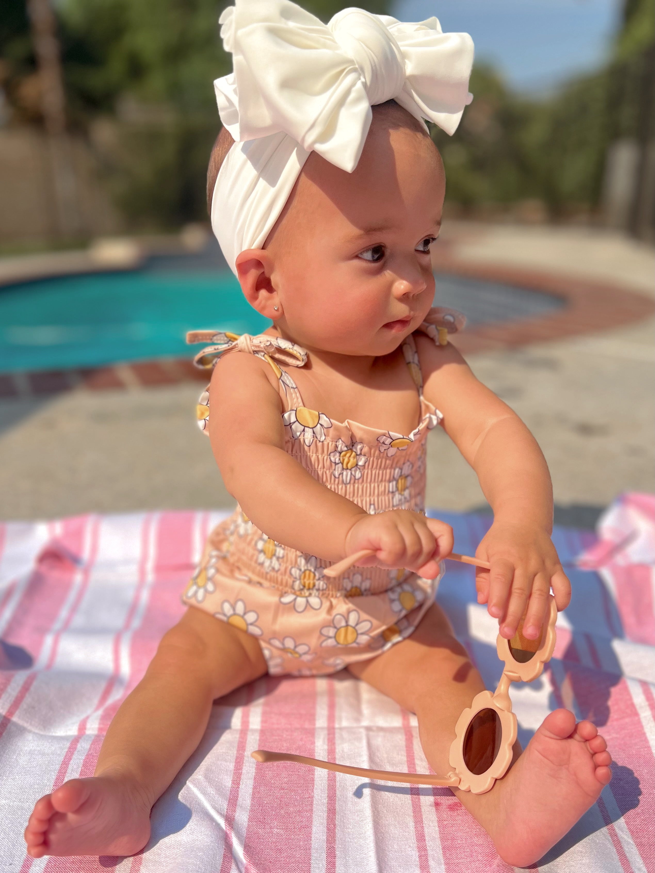 Baby in a floral swimsuit and large bow headband playing with sunglasses on a sunlit towel by a pool.