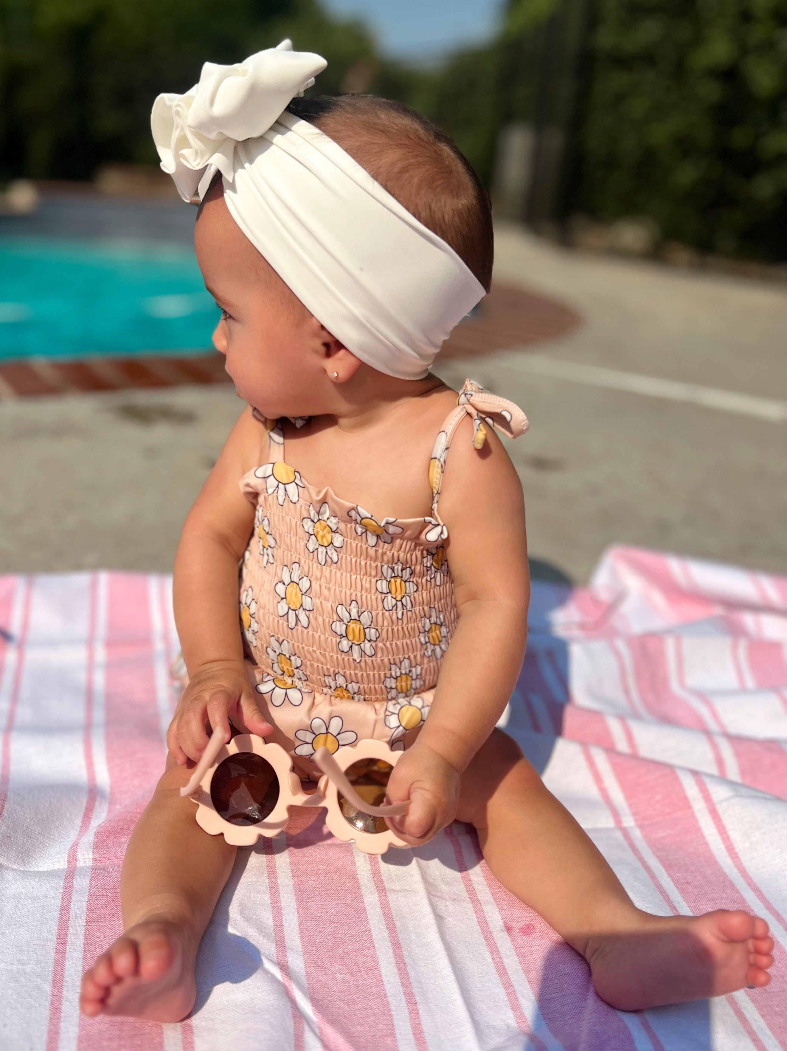 Baby in a floral swimsuit and headband, sitting on a towel by the pool, holding sunglasses.