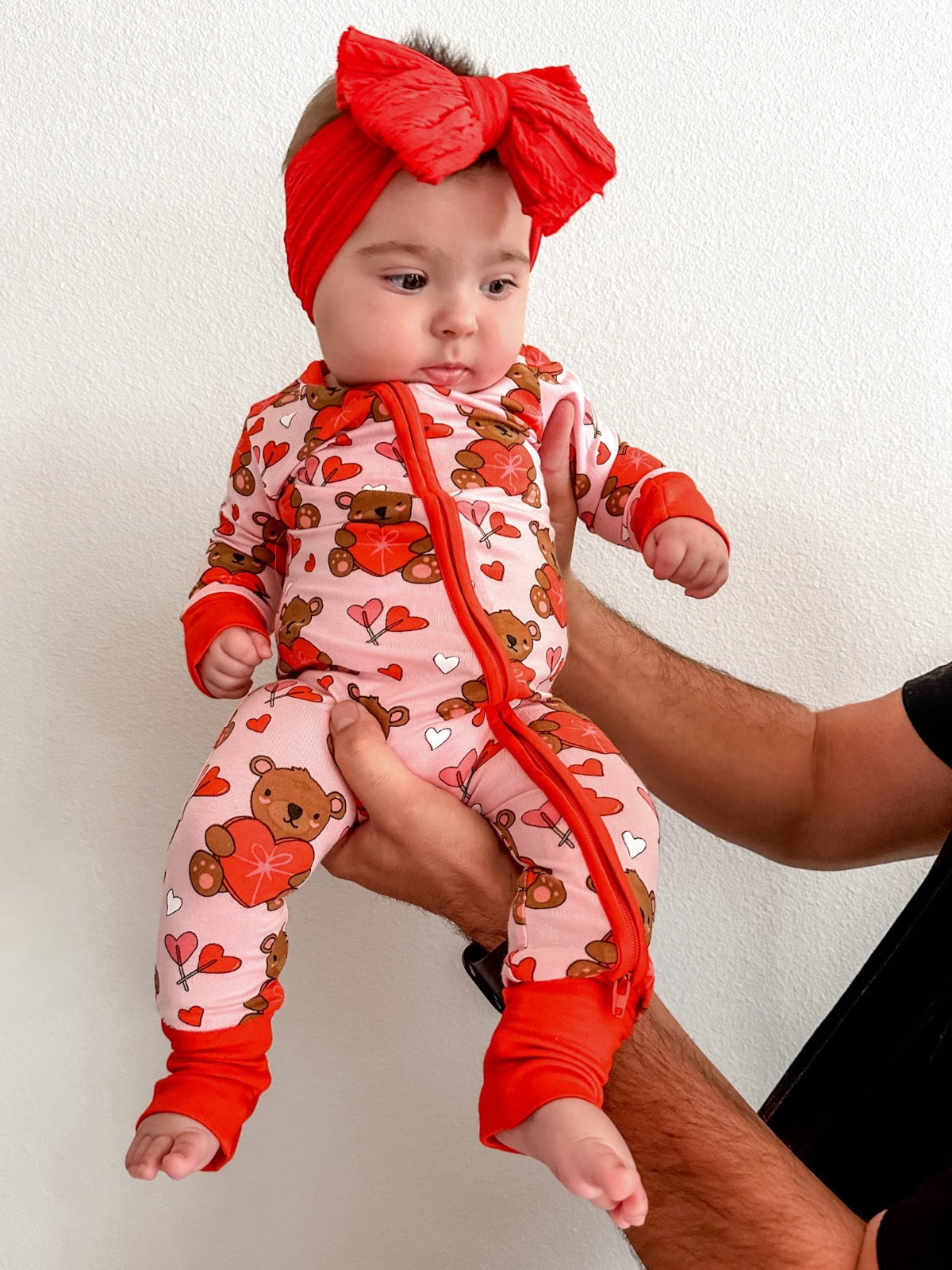 Baby in colorful bear-themed onesie with red accents, held by an adult against a white background.