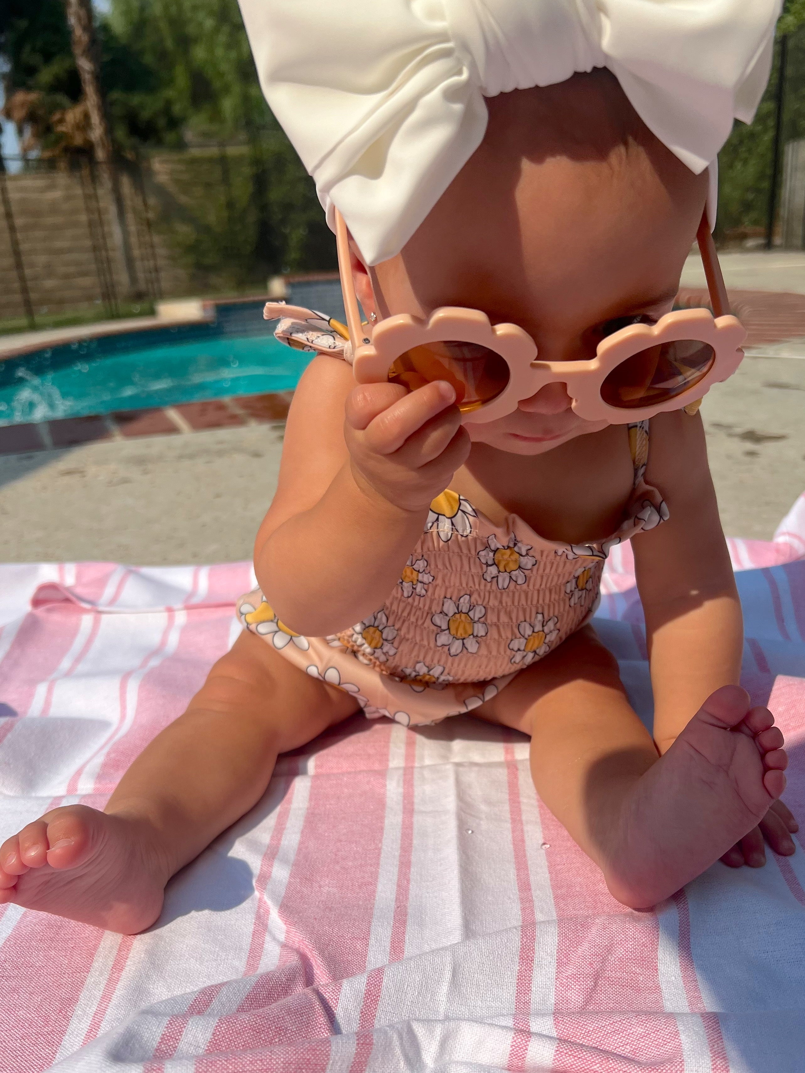 A baby in a floral swimsuit and oversized bow, wearing sunglasses, sits on a beach towel by a pool.