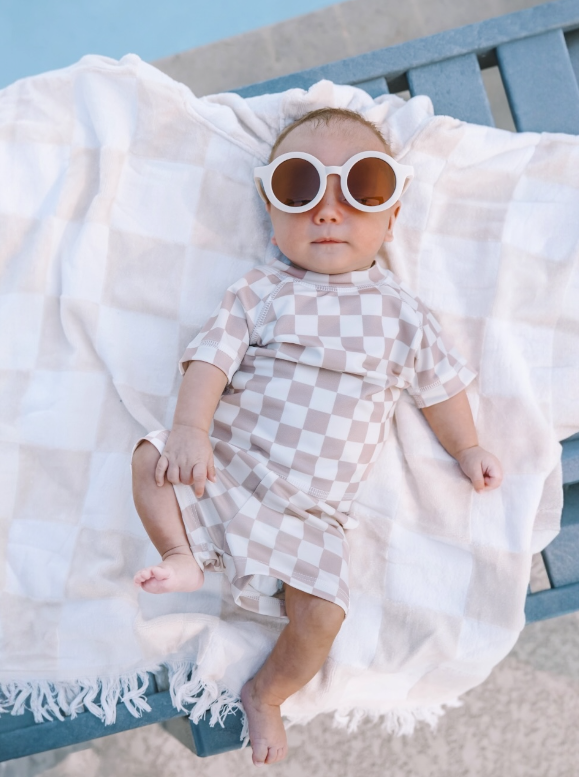 Baby in checkered swim suit and oversized sunglasses lying on a blanket by the pool.