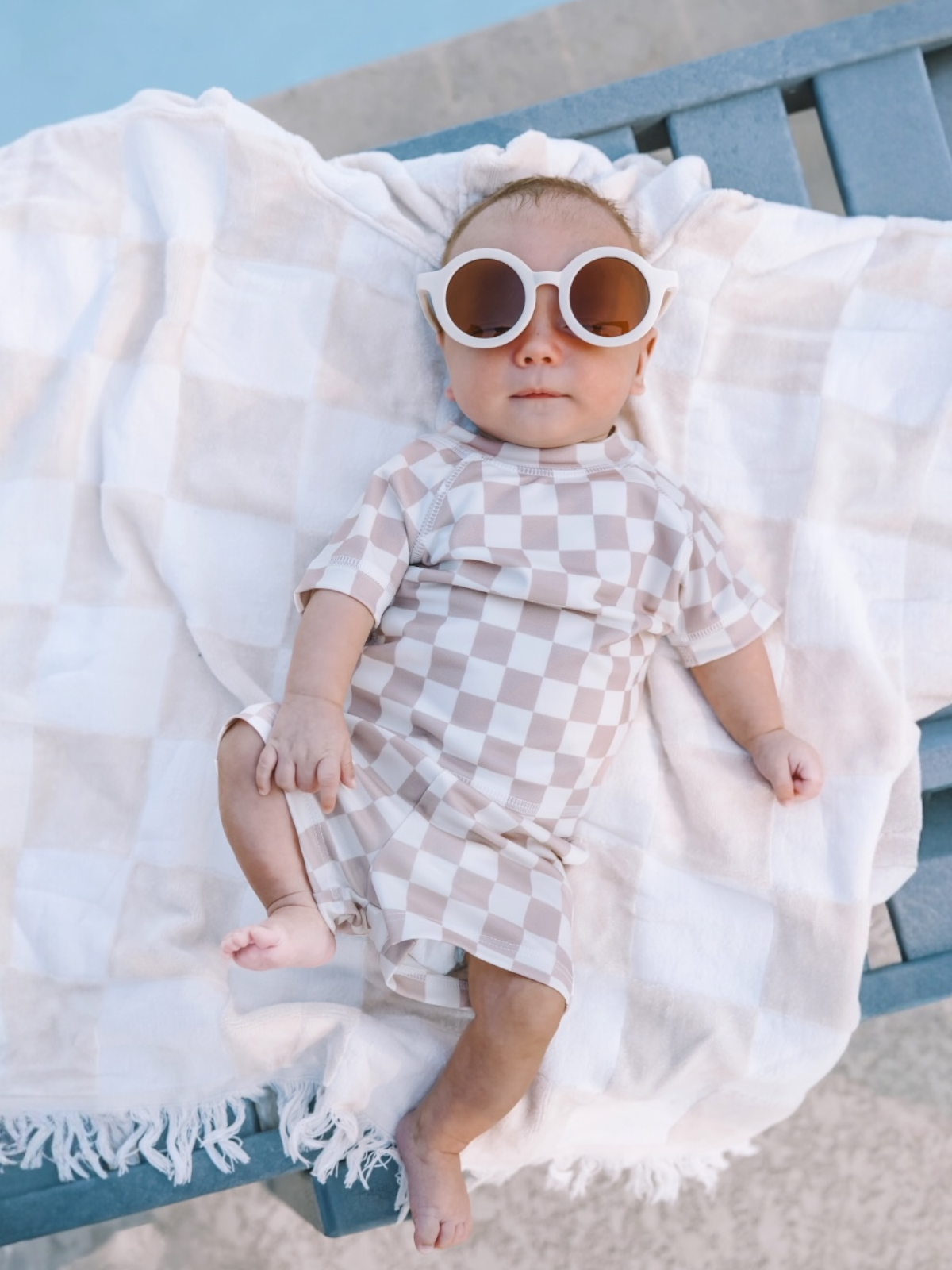Baby in checkered swim suit and oversized sunglasses lying on a blanket by the pool.