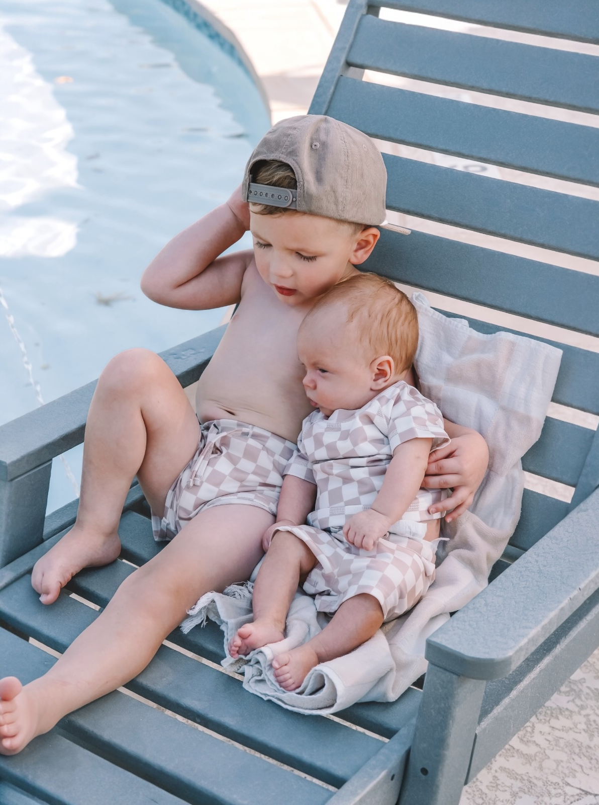 Young boy sits on a poolside chair, holding an infant in matching swimwear, with a pool in the background.