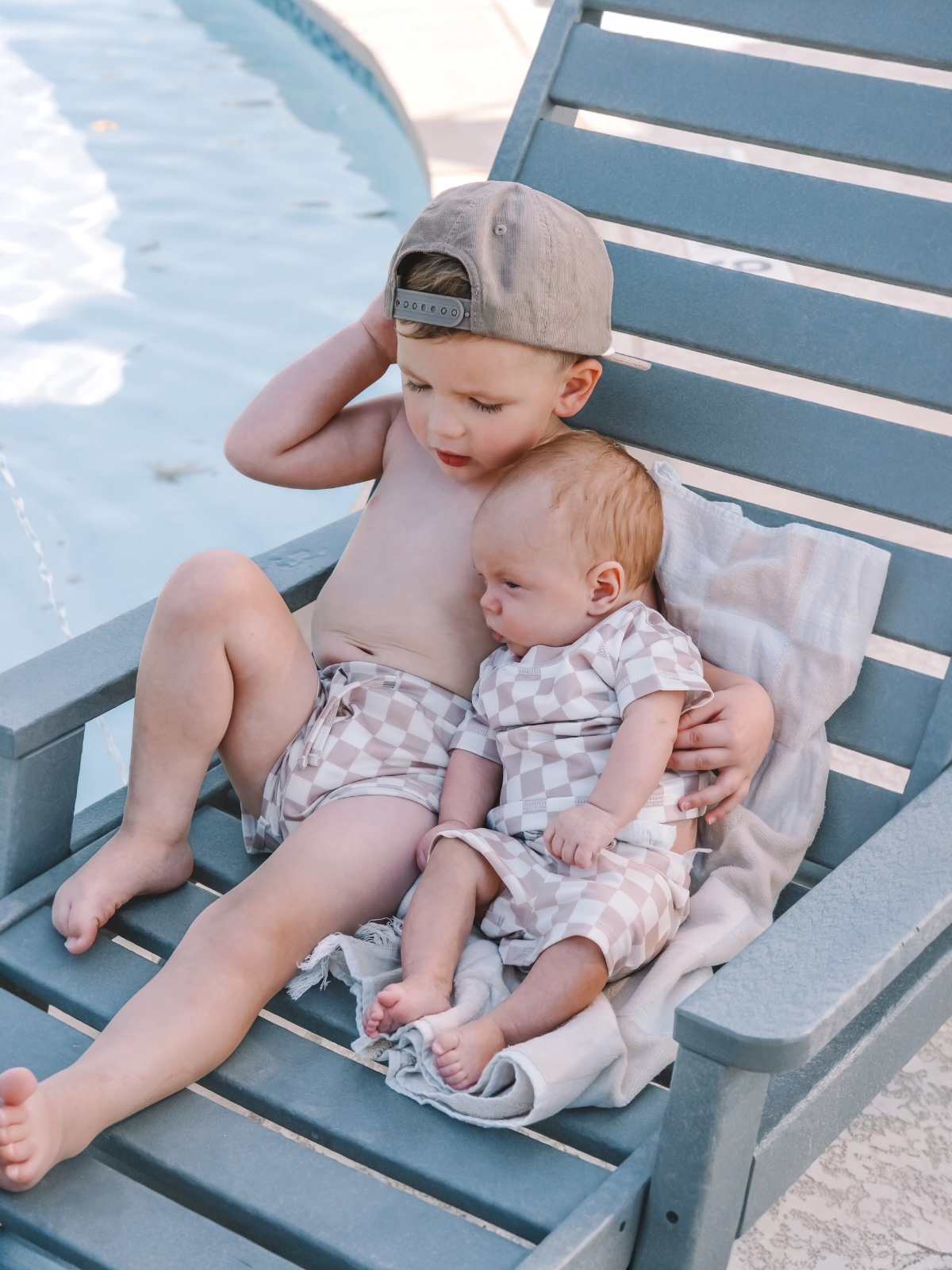 Young boy sits on a poolside chair, holding an infant in matching swimwear, with a pool in the background.