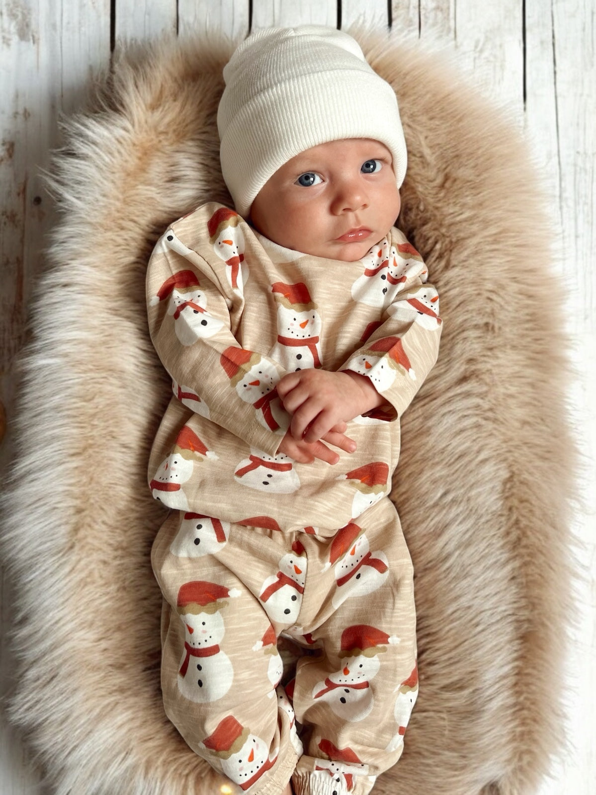 Infant resting on a plush fur blanket, wearing a festive snowman-patterned outfit and a white beanie.