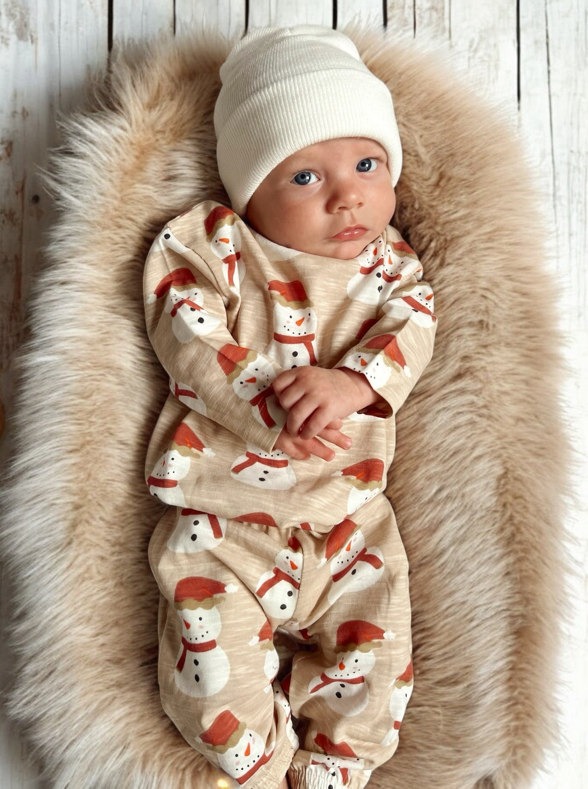 Infant resting on a plush fur blanket, wearing a festive snowman-patterned outfit and a white beanie.