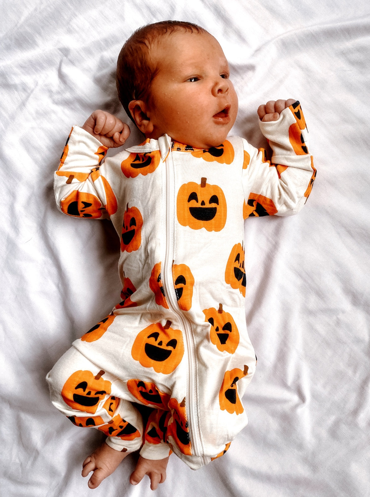 Baby in pumpkin-patterned onesie lying on a white blanket, smiling with hands raised.