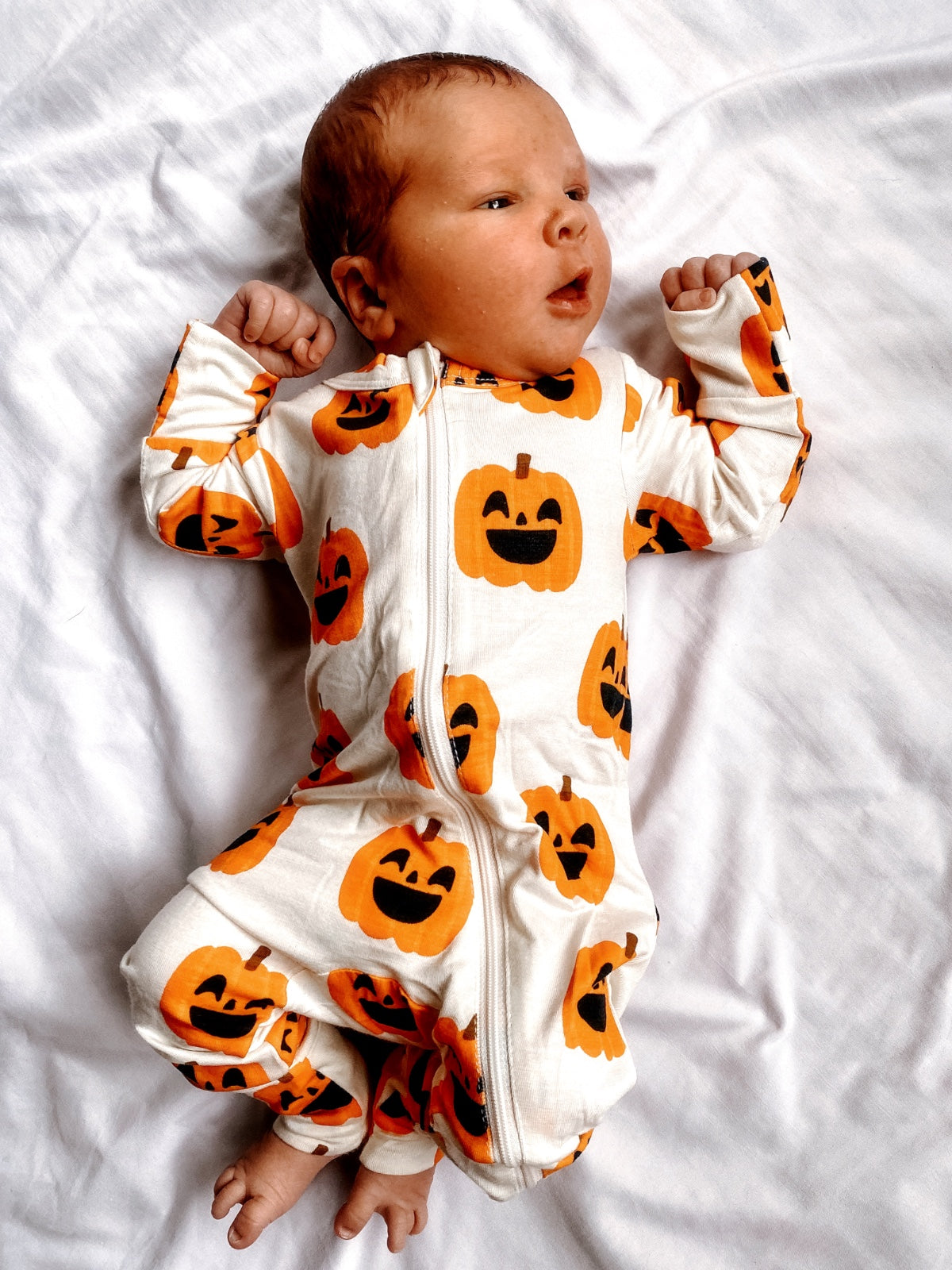 Baby in pumpkin-patterned onesie lying on a white blanket, smiling with hands raised.