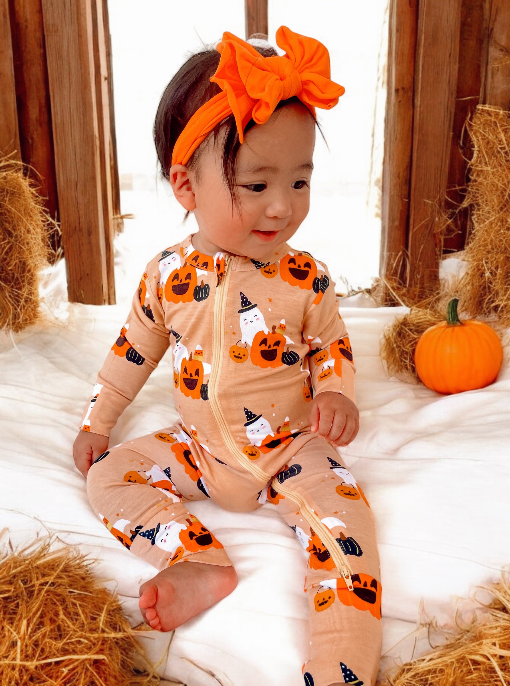 Toddler in Halloween-themed pajamas and orange headband, sitting on white fabric with pumpkins and hay.