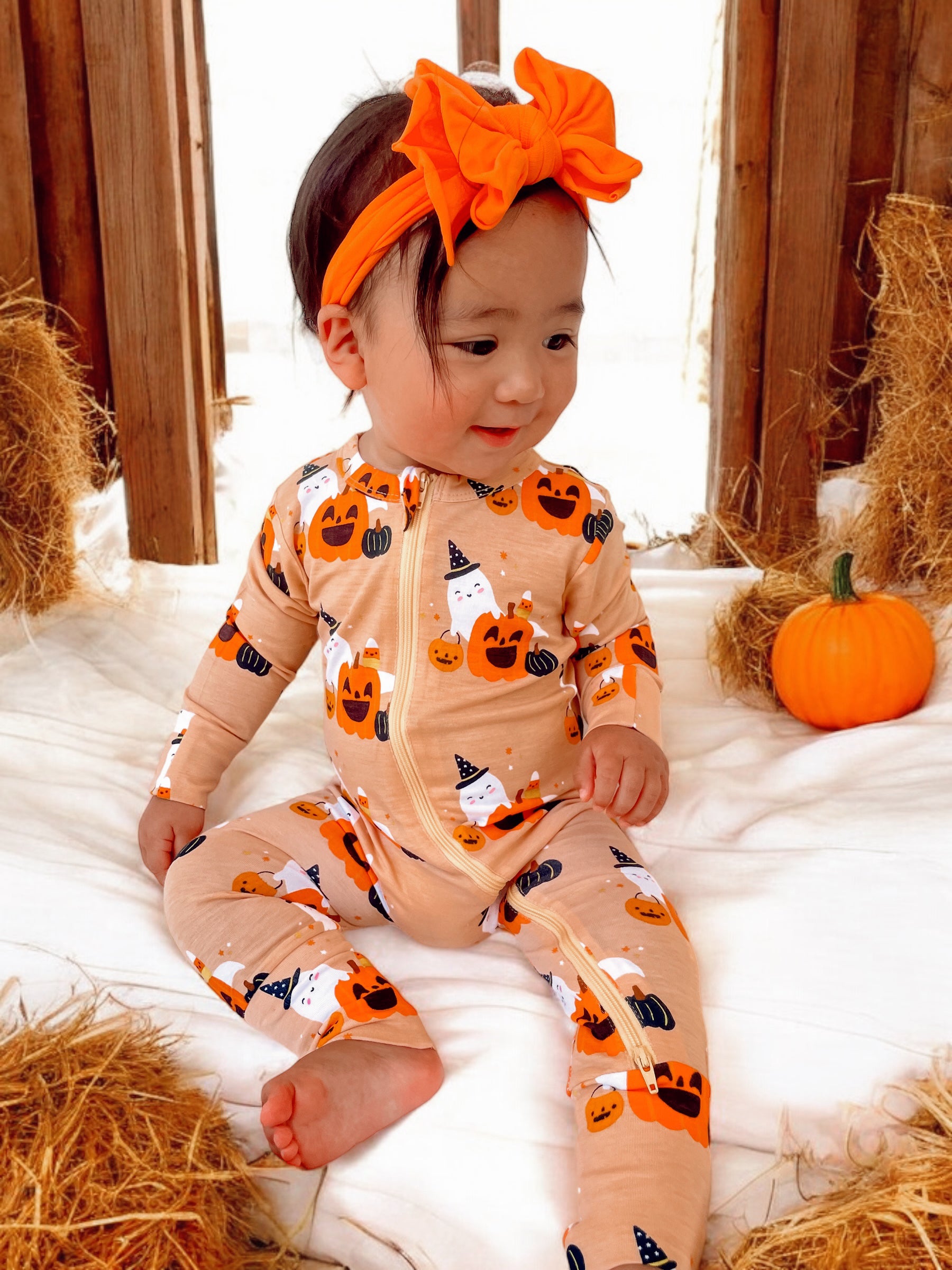 Toddler in Halloween-themed pajamas and orange headband, sitting on white fabric with pumpkins and hay.