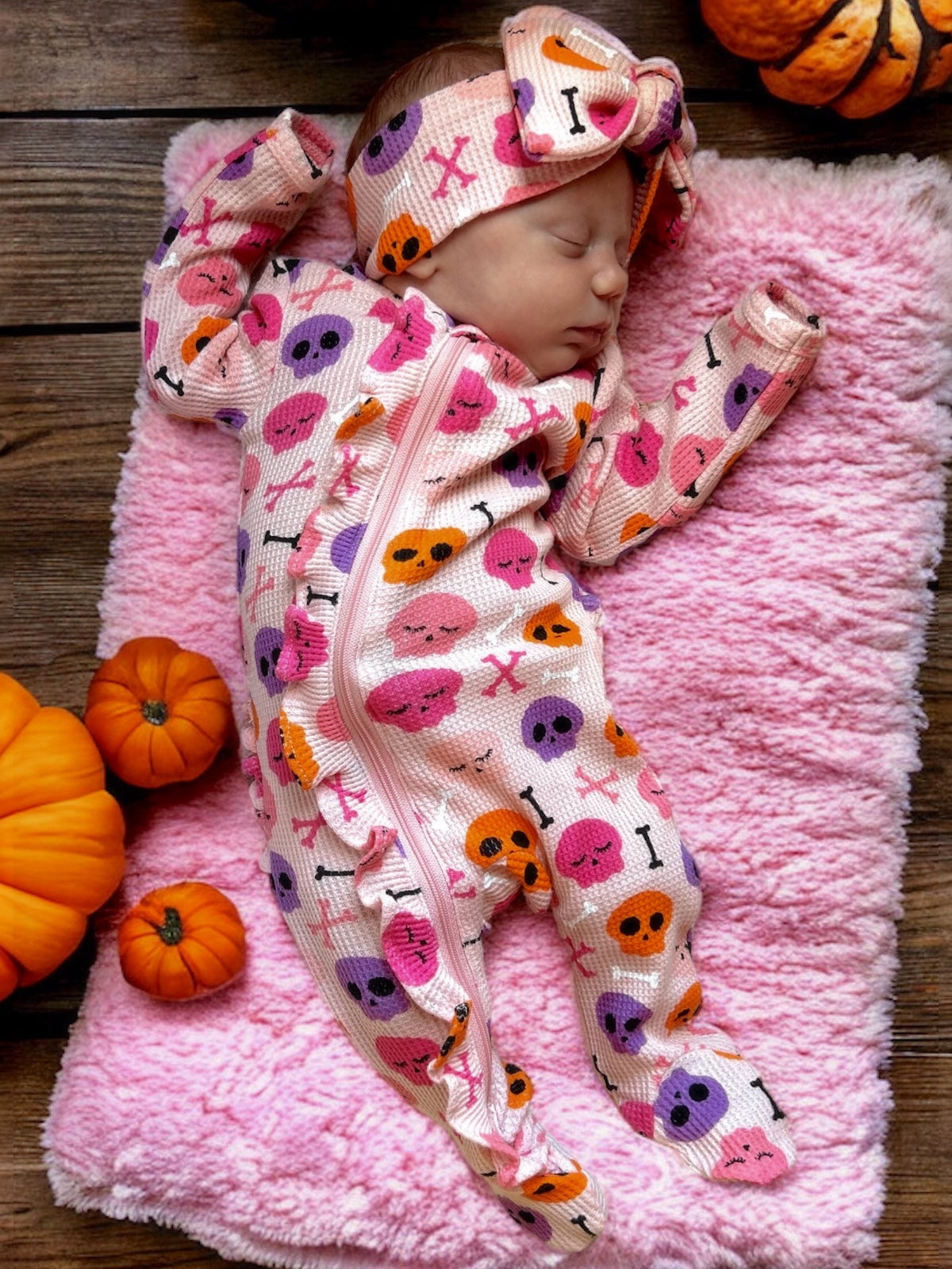 Baby girl in a pink skull-print onesie and matching headband, resting on a fluffy pink blanket with pumpkins nearby.