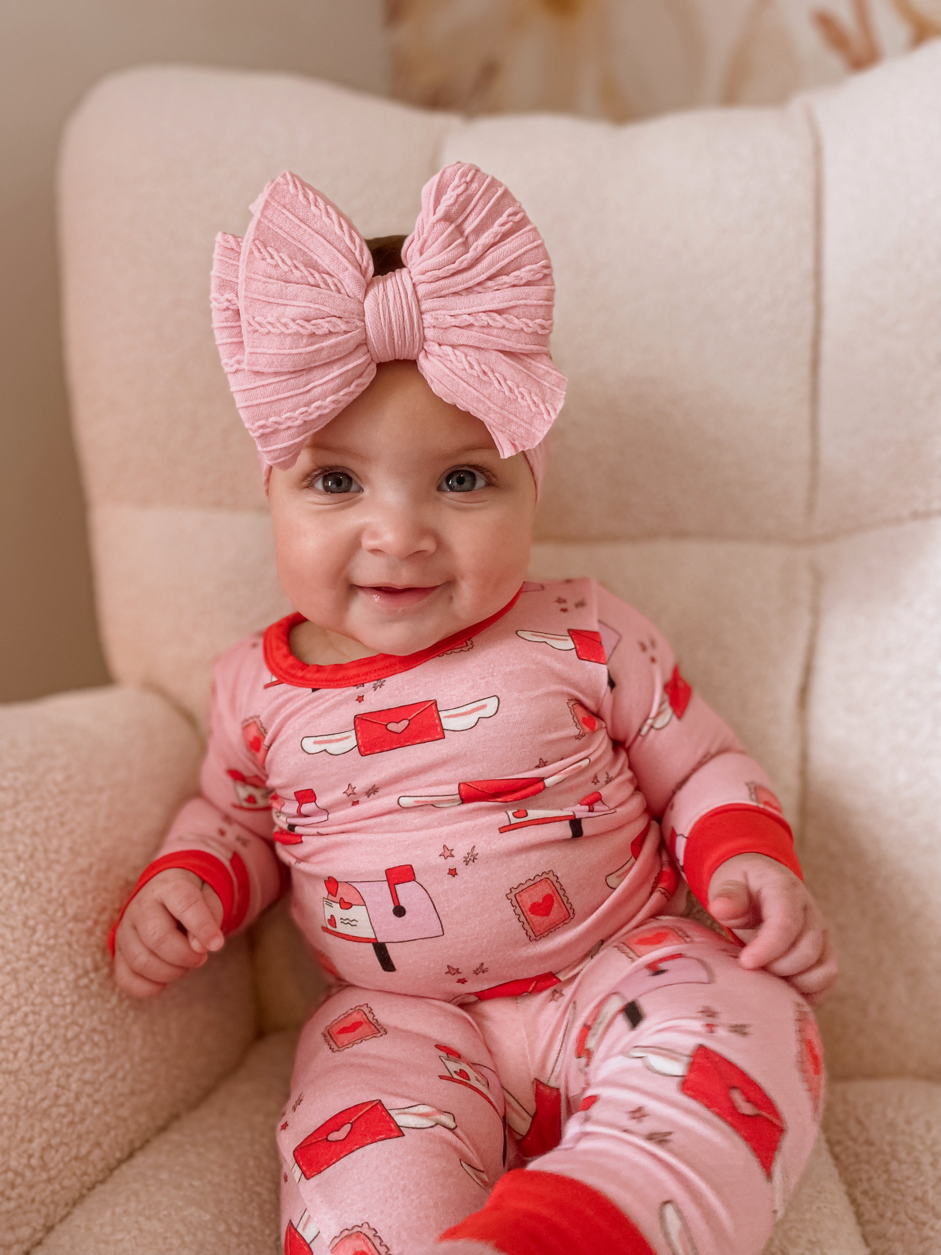 Smiling baby in a pink outfit with a large bow, sitting on a cozy chair.