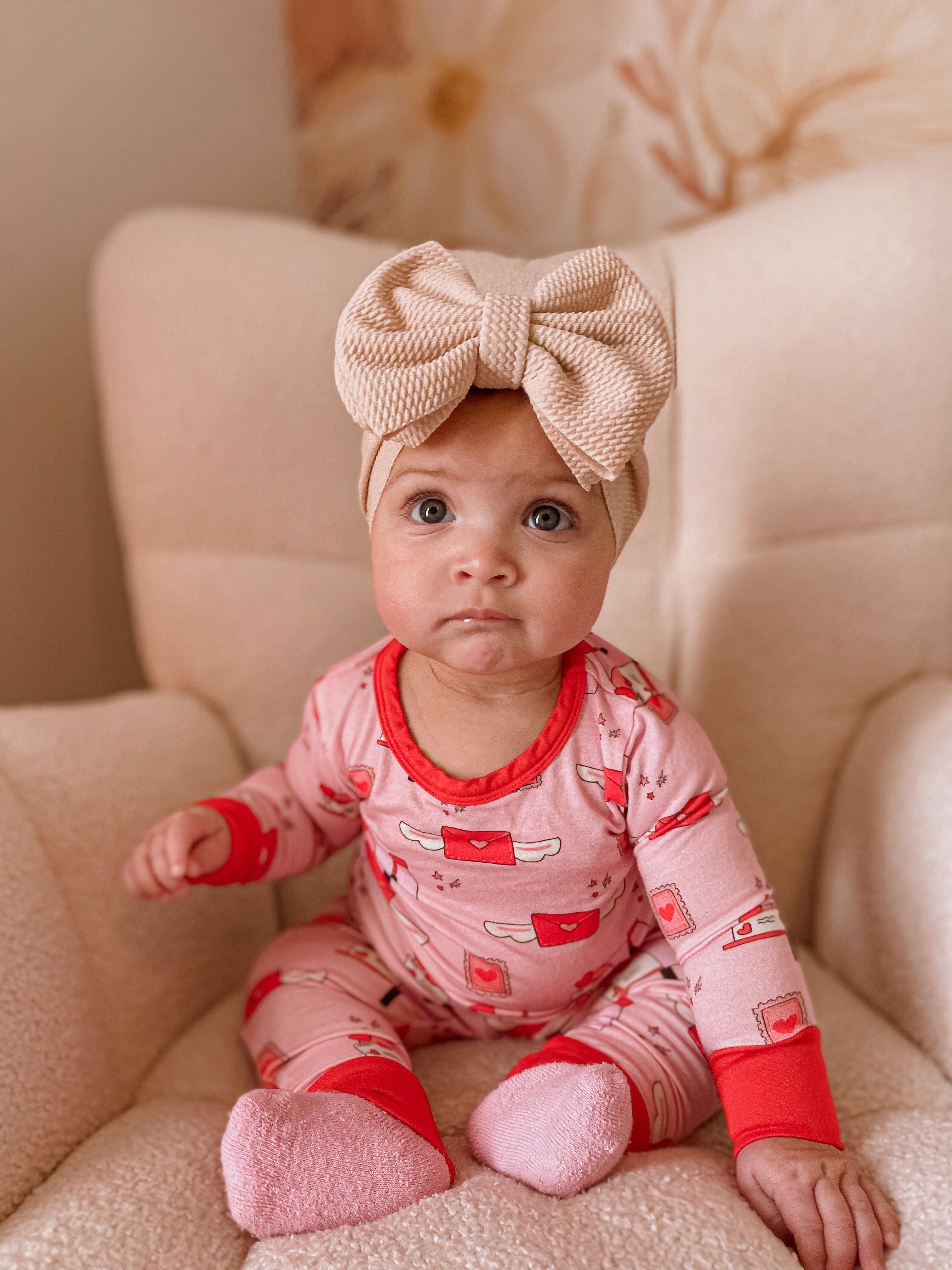 Baby in pink pajamas with red patterns and a large beige bow sits on a cozy chair, looking curiously at the camera.