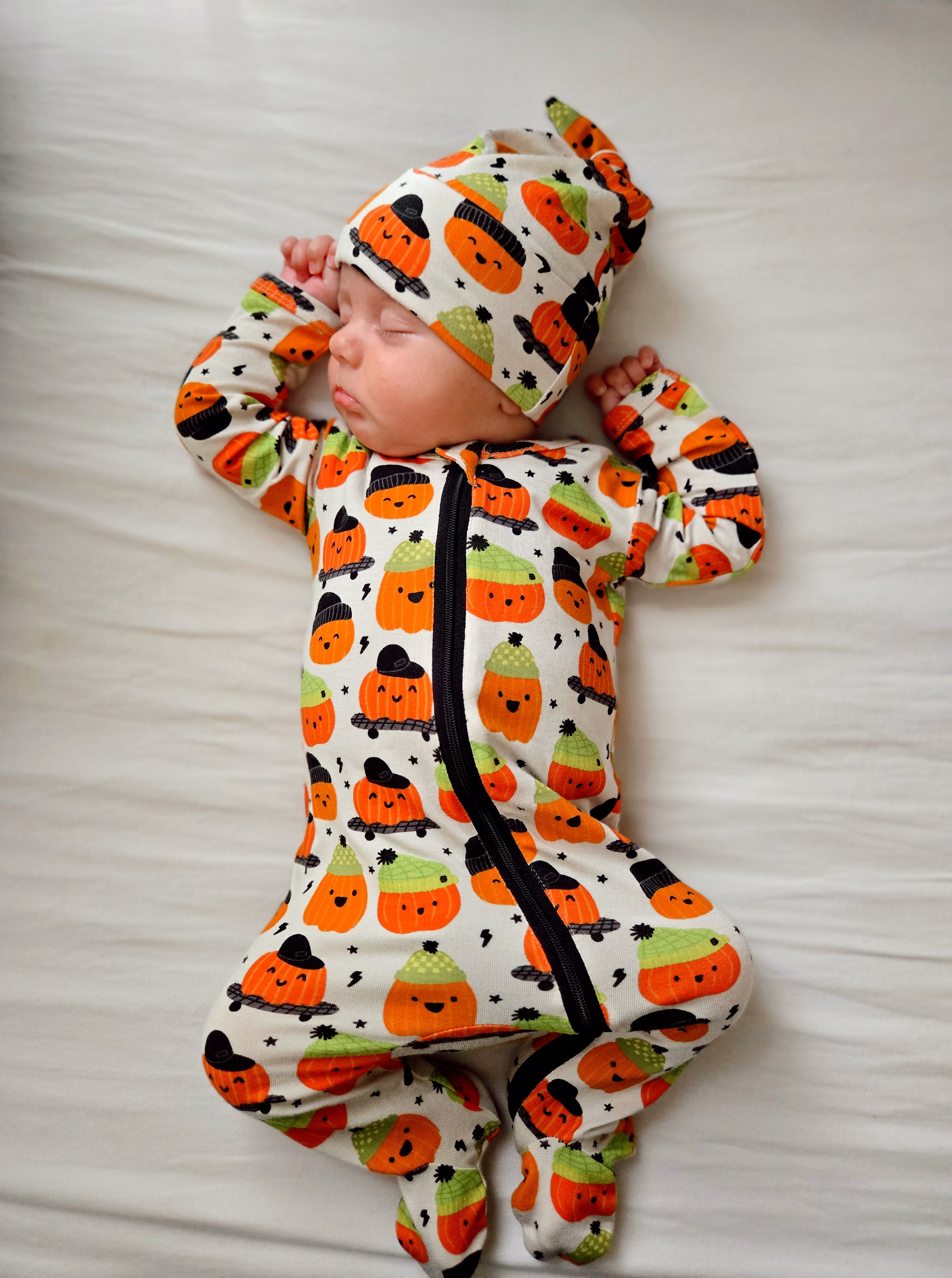 Infant sleeping on a white blanket, wearing a pumpkin-themed outfit and matching hat.
