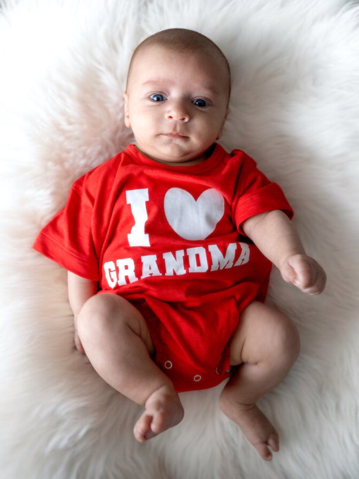 Baby wearing a red "I Love Grandma" shirt, lying on a fluffy white surface. Cute and expressive gaze.