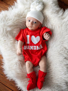 Baby in a red onesie reading "I ❤️ Grandma," wearing a white hat and red booties, on a fluffy white blanket.