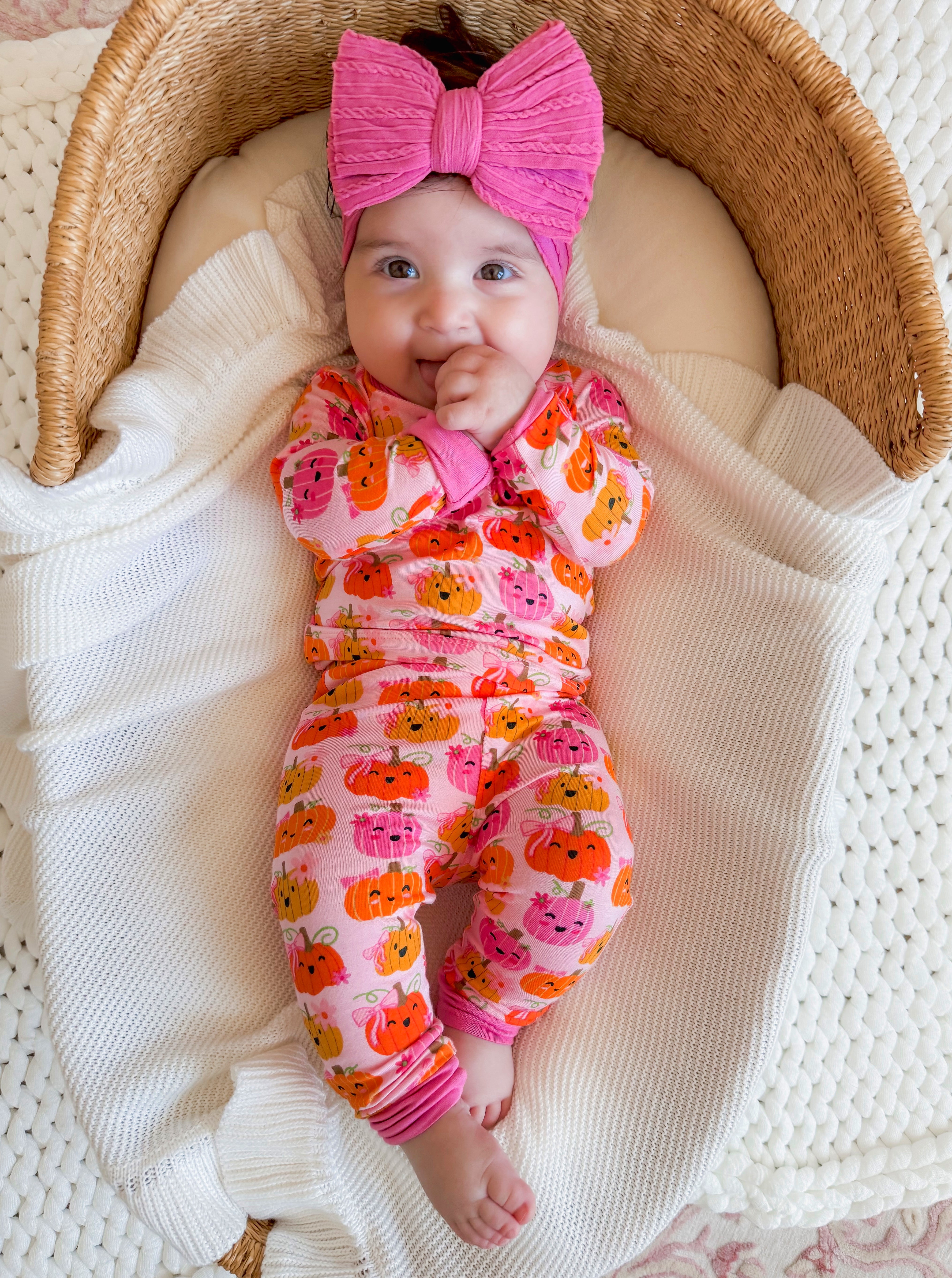 Smiling baby in a pumpkin-patterned outfit, wearing a large pink bow, nestled in a cozy woven basket.