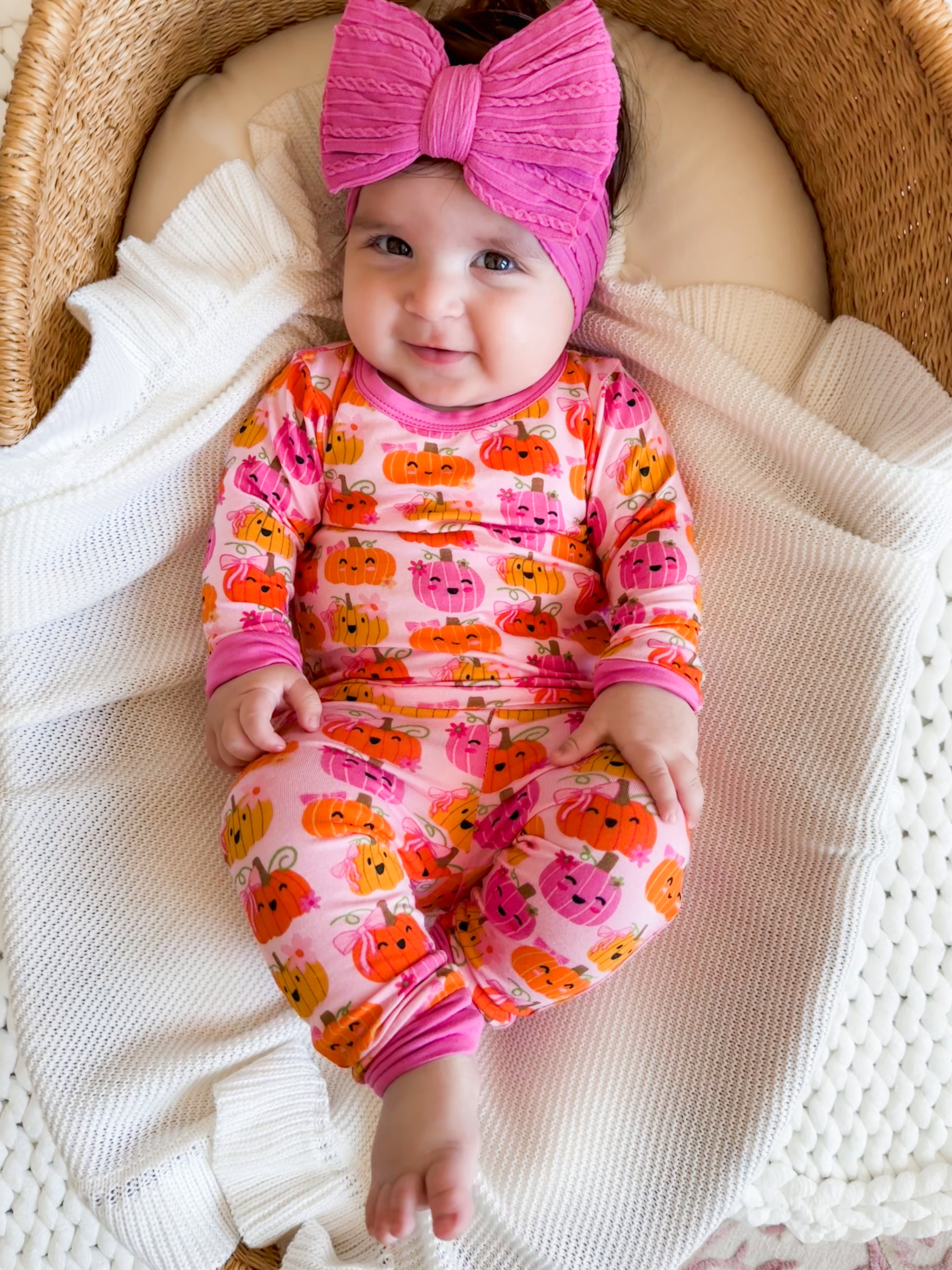 Smiling baby in a pink pumpkin-print outfit and a matching headband, sitting on a cozy blanket in a woven basket.